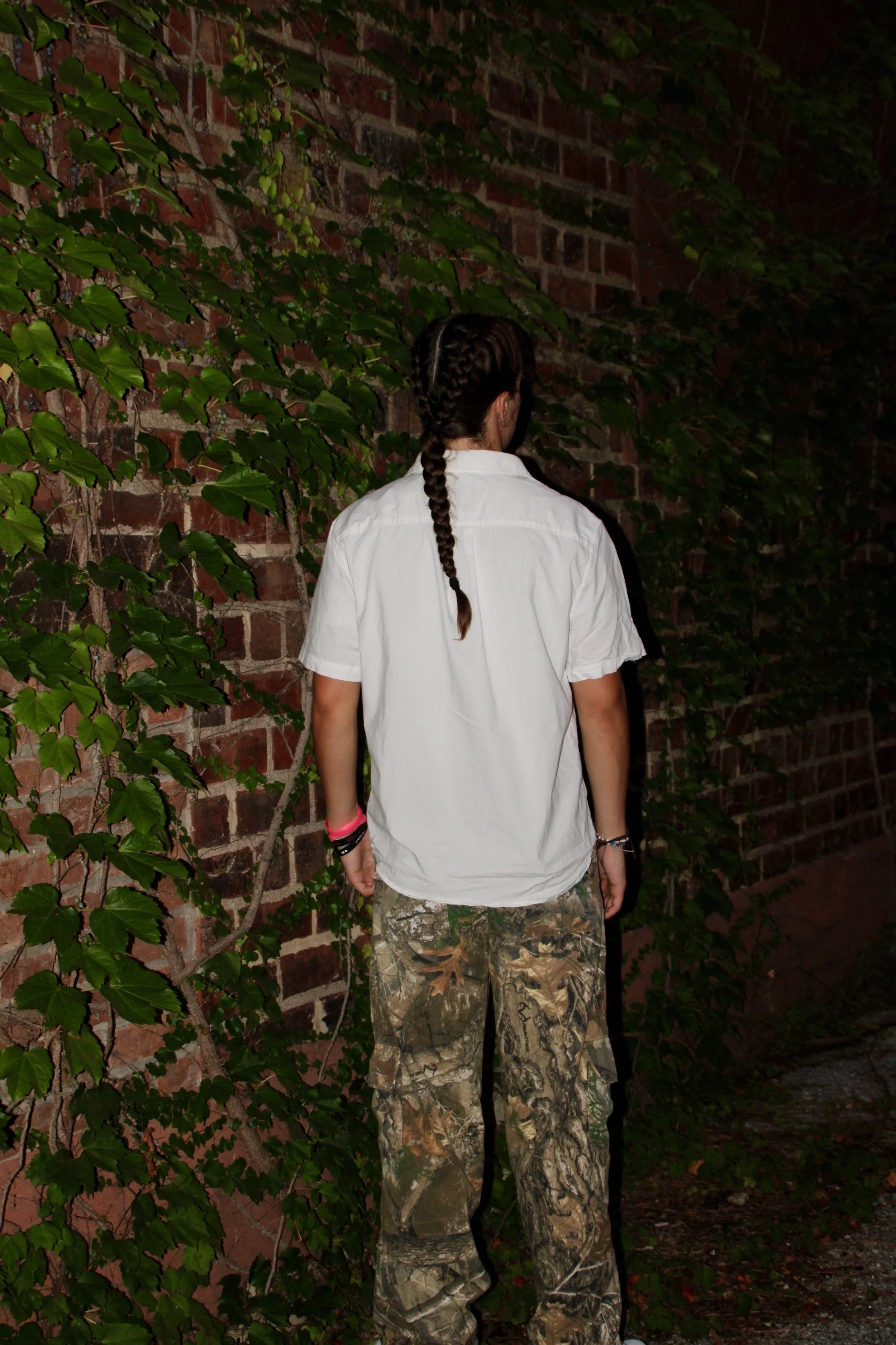 A woman with long hair styled in multiple braids, wearing a white shirt and camouflage pants, standing with her back facing the camera near a brick wall covered in green ivy, at night.