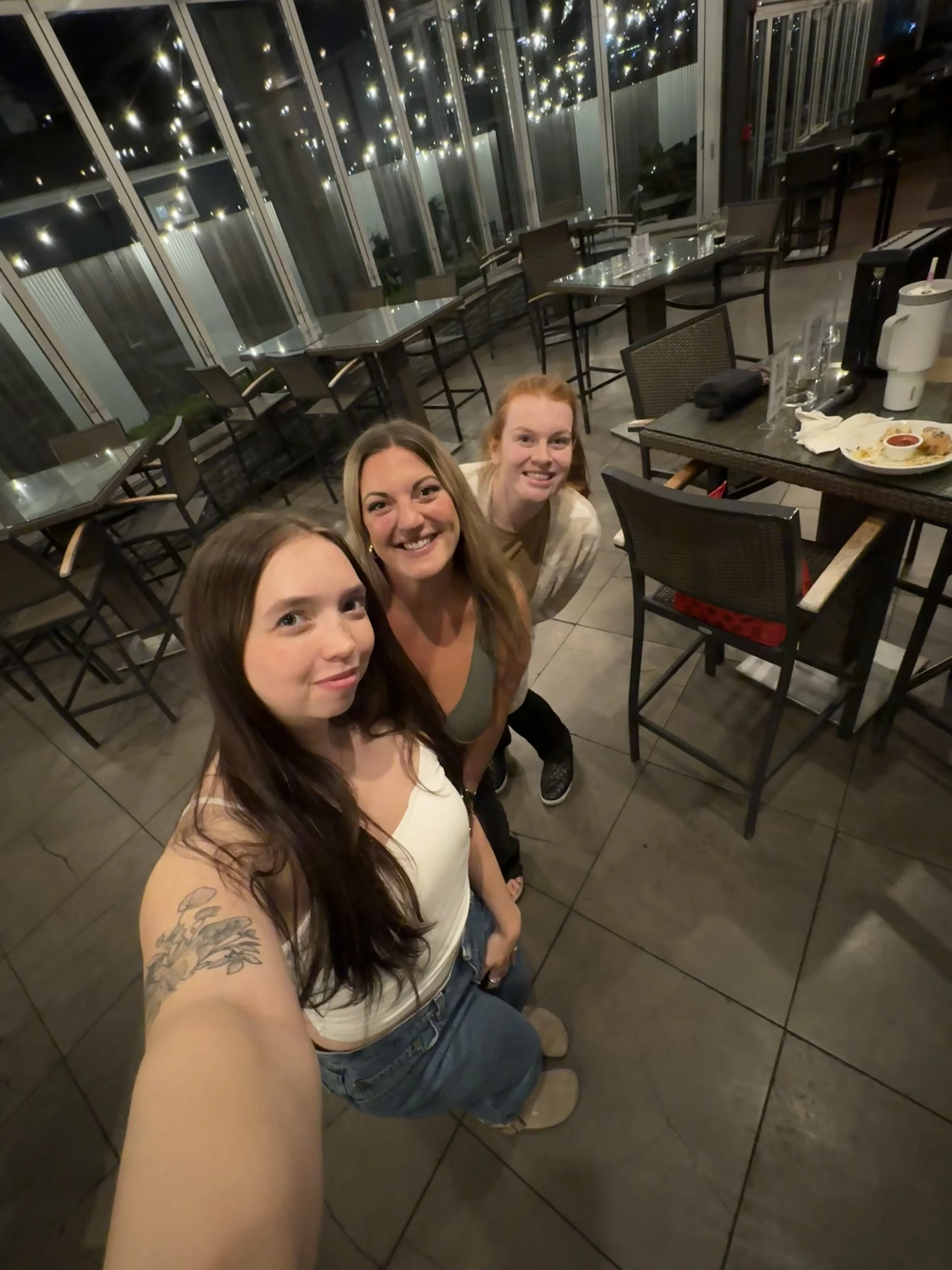 Three women are smiling for a selfie in a restaurant with large windows reflecting lights.