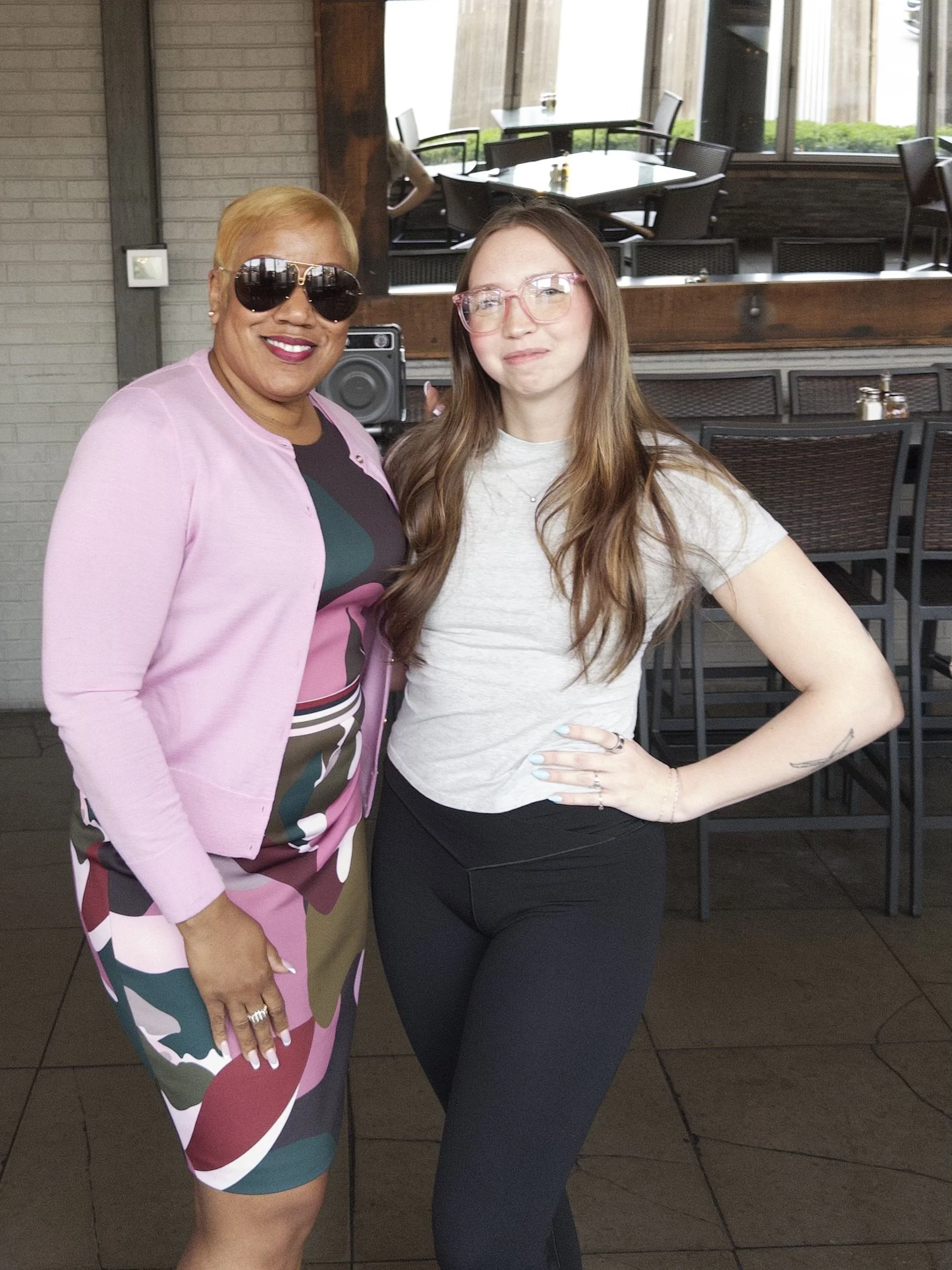 Two women are standing indoors, one wearing sunglasses and a colorful dress with a pink blazer, the other with long brown hair, glasses, a light gray T-shirt, and black pants, smiling for the photo.