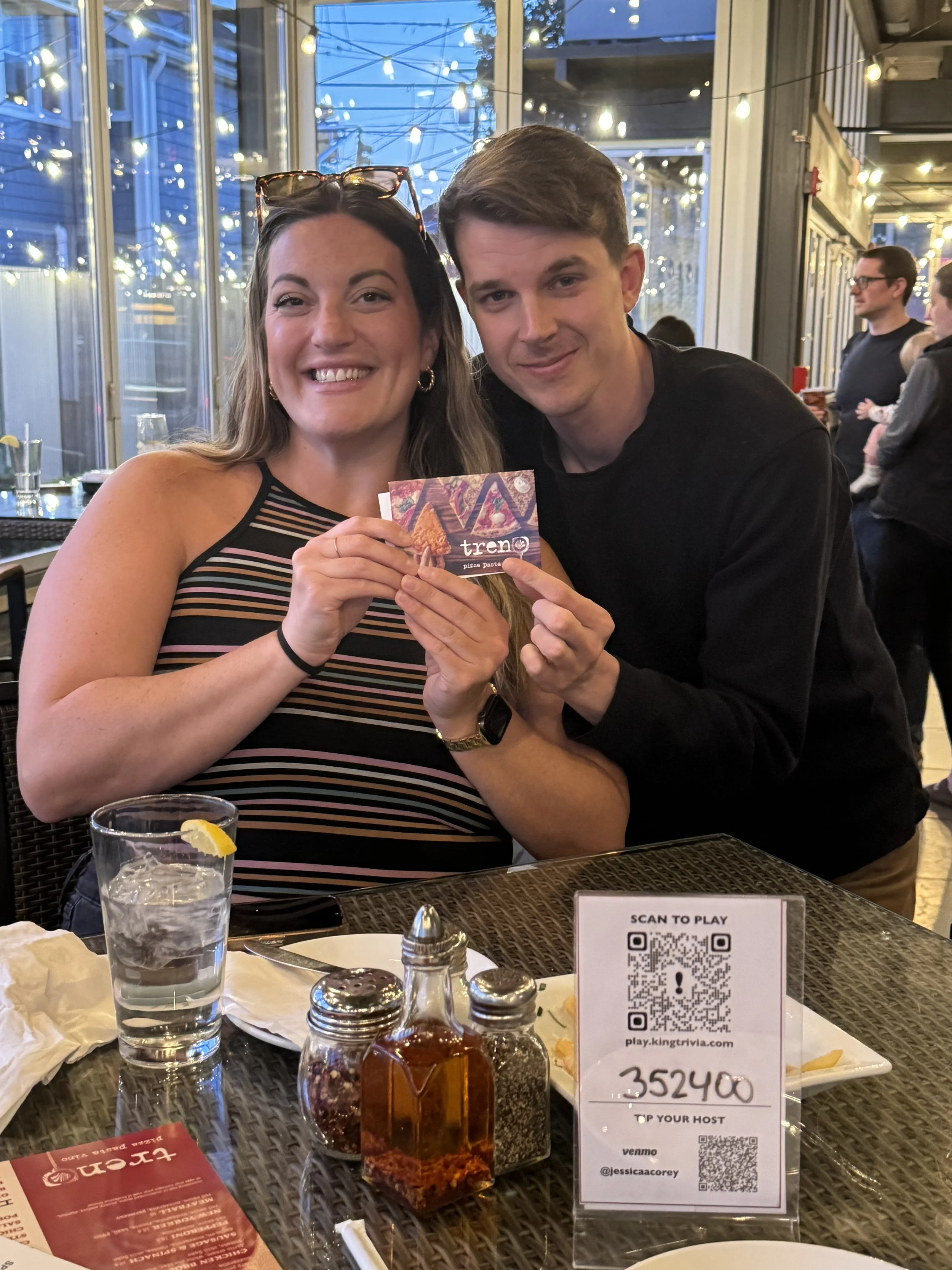 A woman and a man smiling and holding a small card at a restaurant. The woman is wearing a striped top and sunglasses on her head, while the man is in a black shirt. Table items include a glass of water with lemon, condiments, and a sign with a QR co