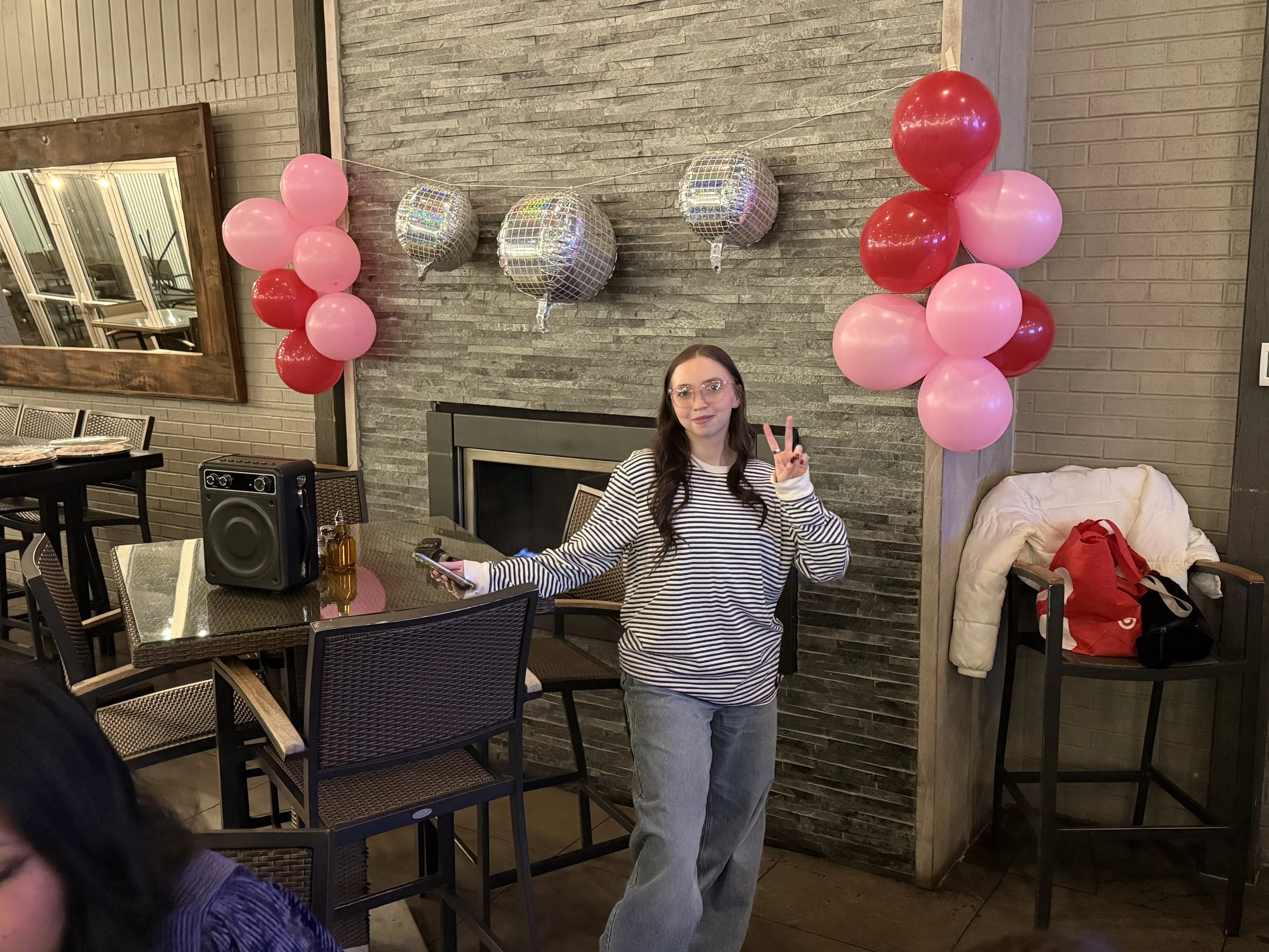 A woman with long dark hair and glasses, wearing a black and white striped shirt, making a peace sign with her right hand, sitting in a restaurant or event space with pink and red balloons and disco ball balloons in the background.