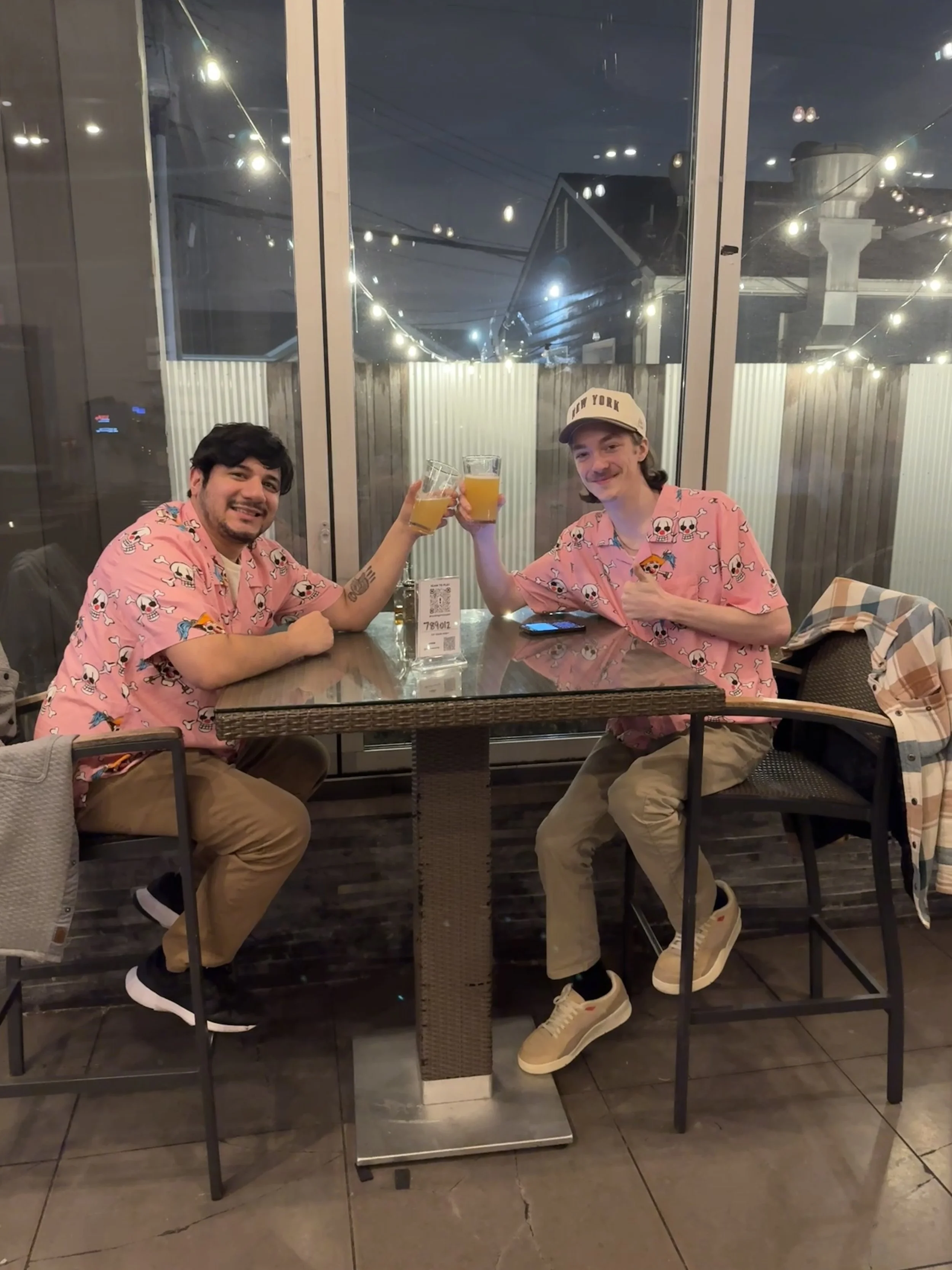 Two young men in pink skull-printed shirts are sitting at a table, smiling, and clinking glasses of beer inside a restaurant with a large glass window and string lights outside.