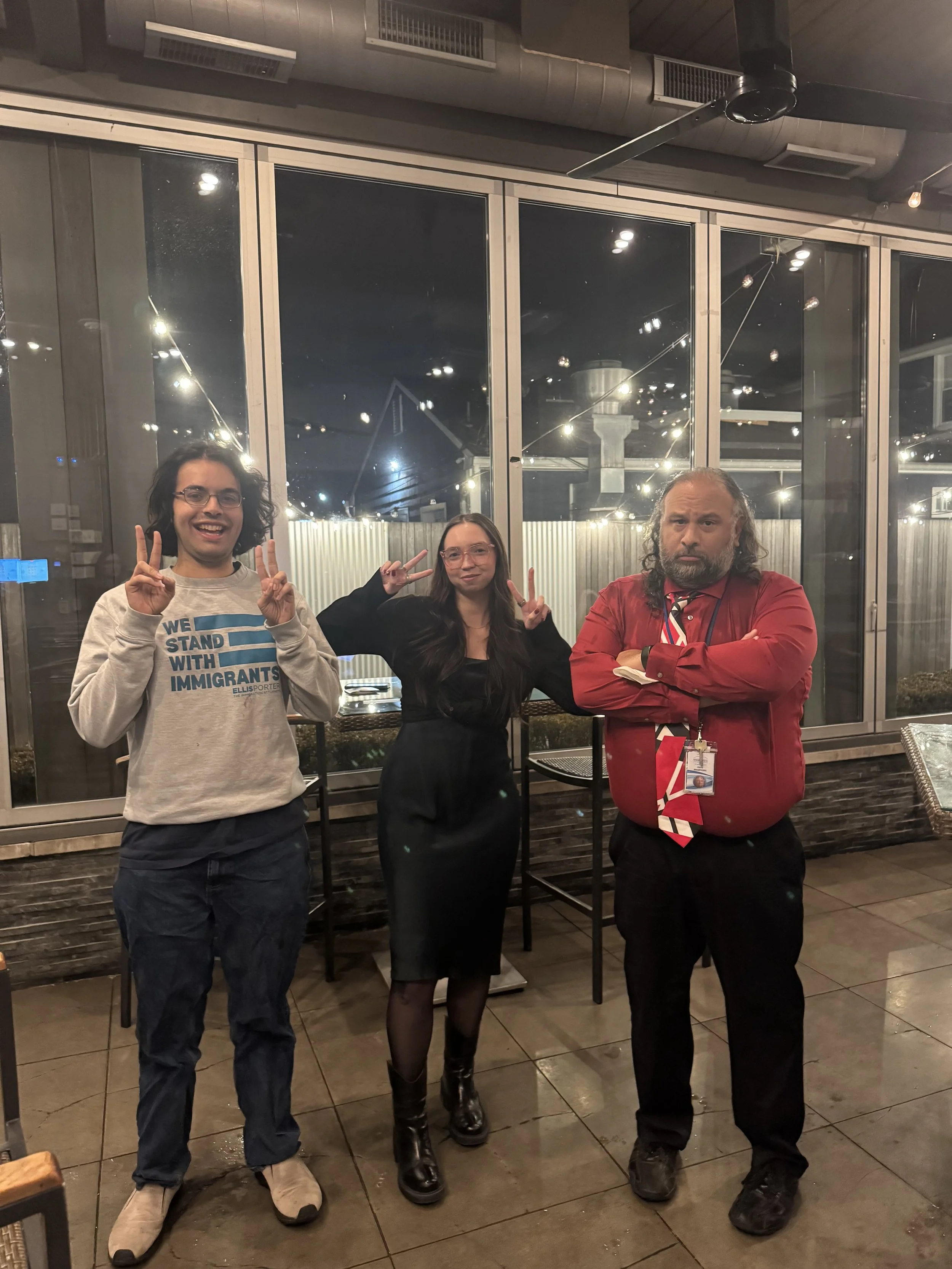 Three people standing indoors, posing for the camera. The person on the left is a man with long dark curly hair, glasses, a light-colored sweatshirt with text, and dark pants. The person in the middle is a woman with long dark hair, glasses, dressed 