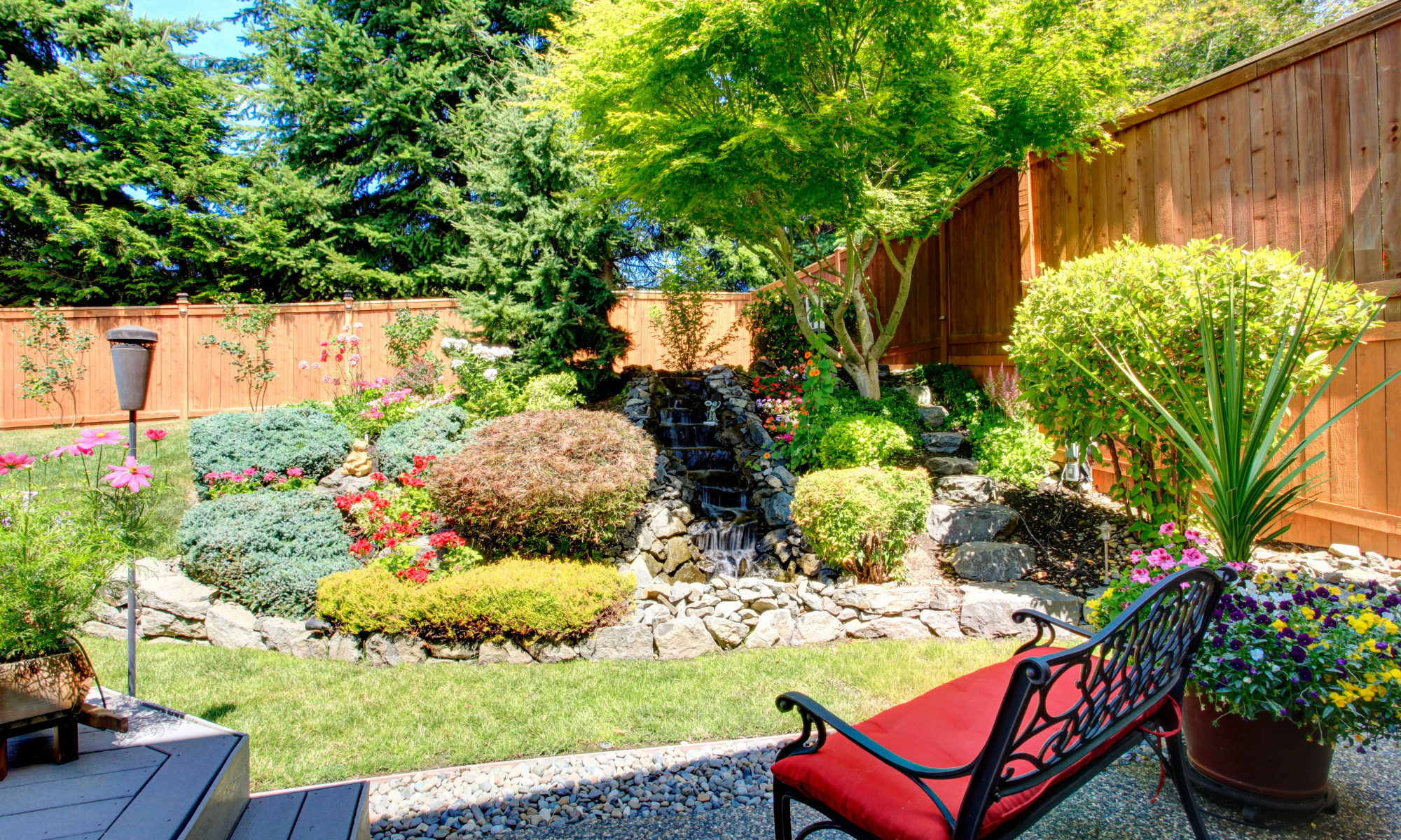 A lush backyard garden with a variety of vibrant flowers, trees, and a small waterfall flowing over rocks. There is a wooden fence surrounding the yard, and a metal bench with a red cushion in the foreground.