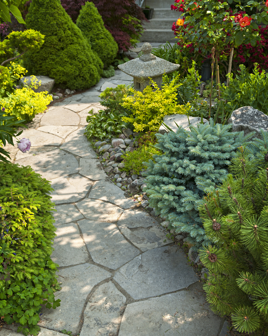 A garden pathway made of irregular stone slabs, surrounded by lush green shrubs and colorful flowering plants, with a small stone lantern near the steps in the background.