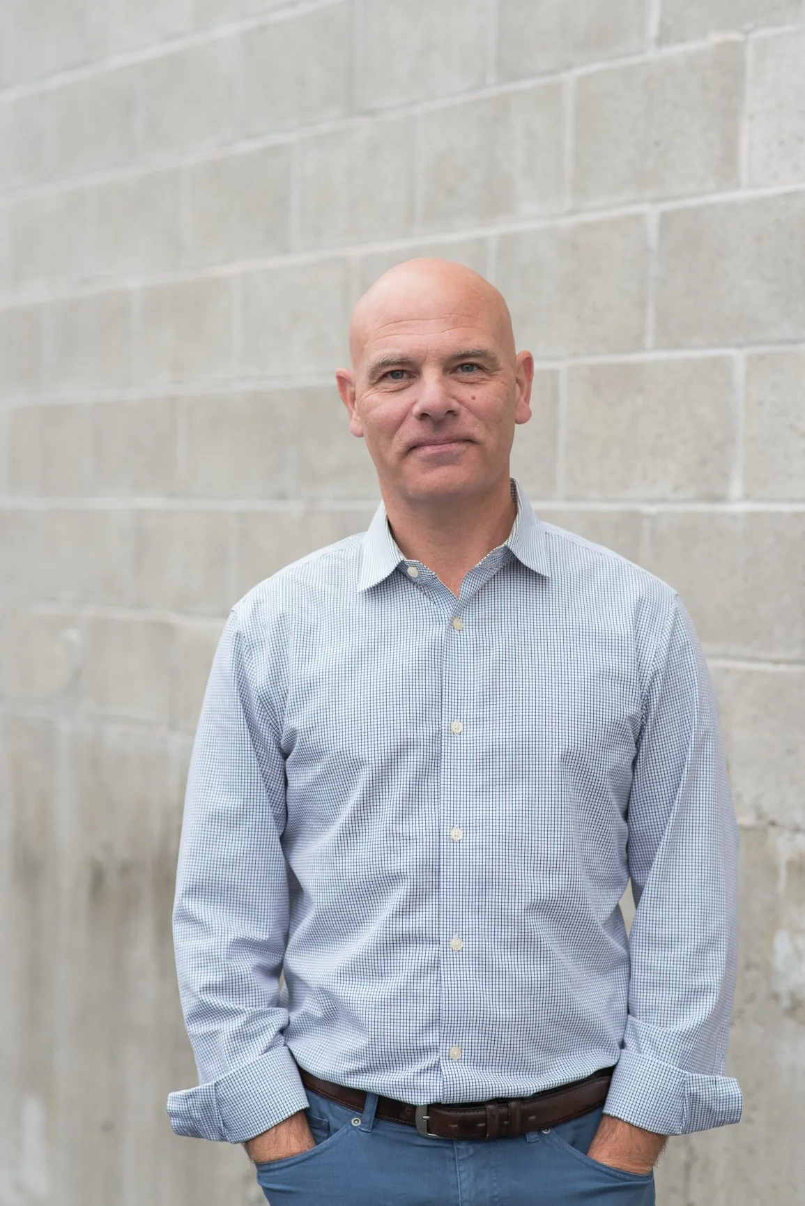 A bald man wearing a light blue checkered button-up shirt and dark pants, standing in front of a beige brick wall with his hands in his pockets, looking at the camera with a slight smile.