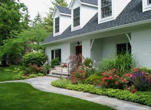 Front view of a house with a garden and walkway, featuring white siding, black shutters, and a well-maintained yard.