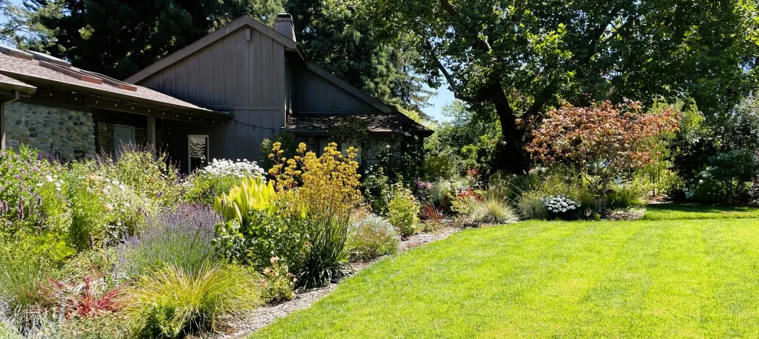 A lush garden with colorful flowers and green grass in front of a house with stone and wooden exterior, surrounded by trees.