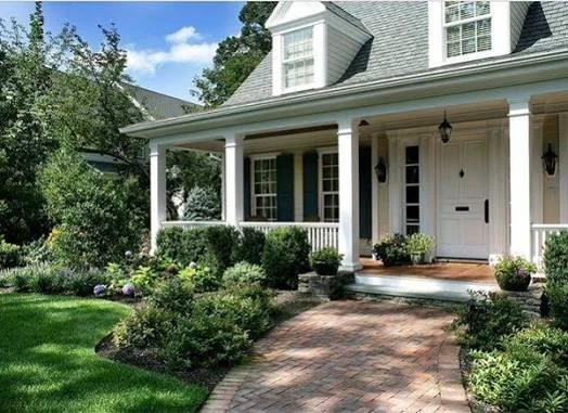Front view of a white house with a porch, brick pathway, and landscaped garden with bushes and flowers.