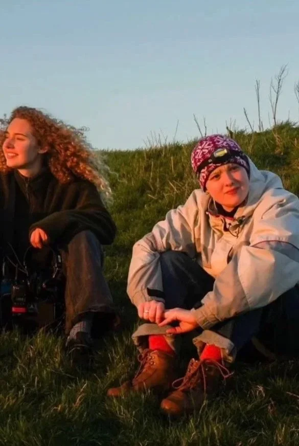 Two women sitting on grassy hill outdoors during sunset, one smiling and the other looking at the camera with a neutral expression, both dressed in outdoor clothing.