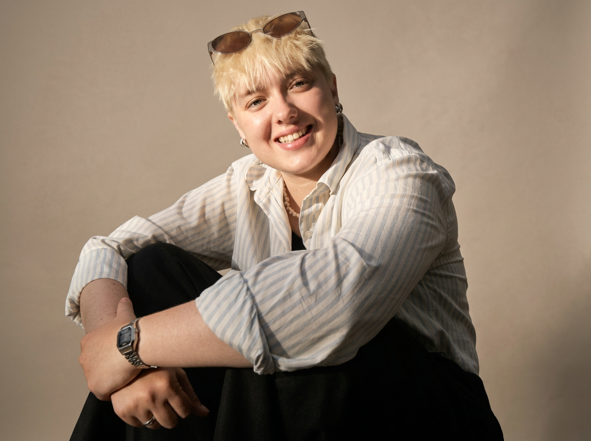 Young woman with short blonde hair, sunglasses on her head, wearing a striped button-up shirt, smiling, sitting with her arms wrapped around her knee.