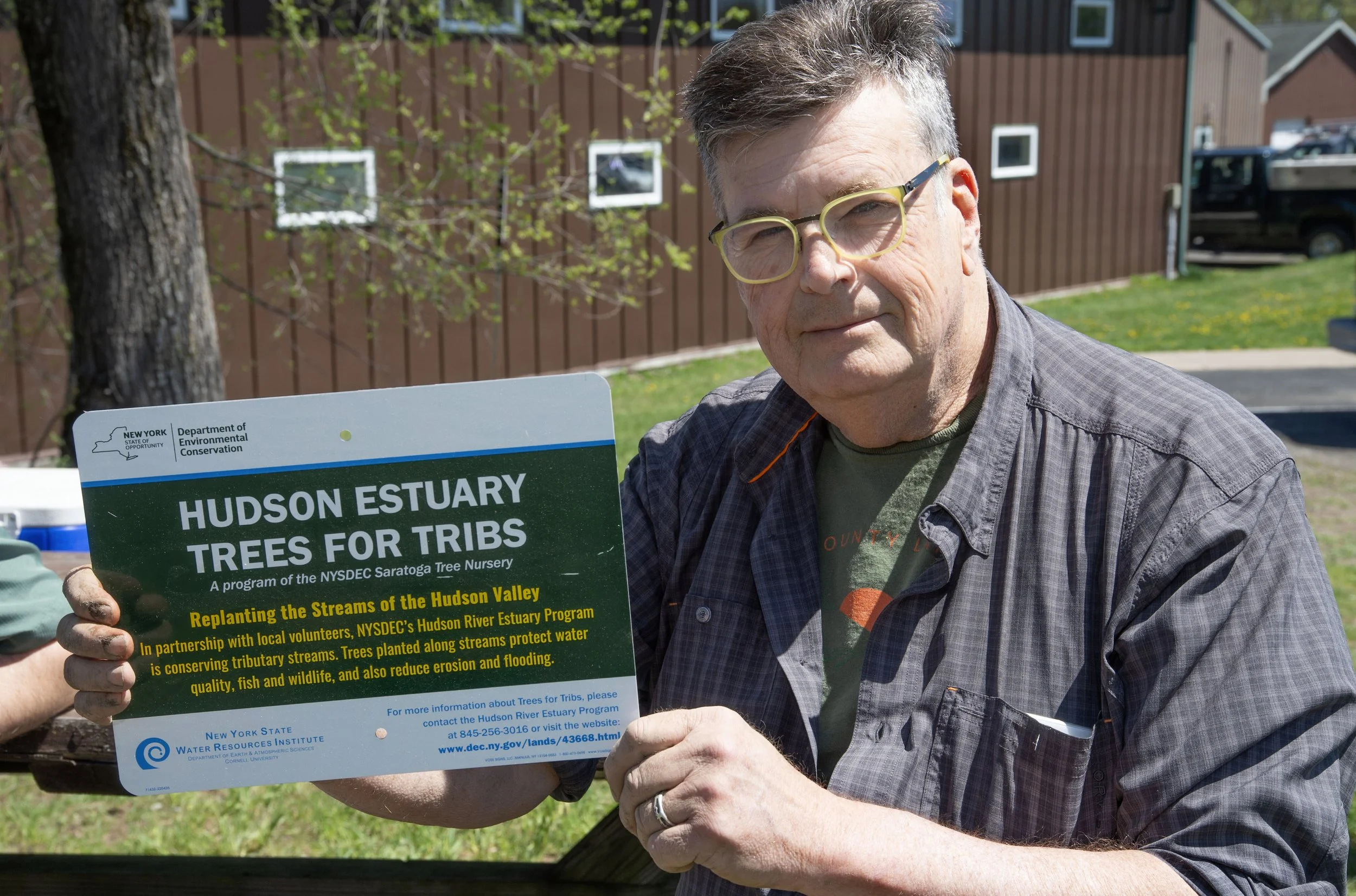 A man with glasses and gray hair holding a sign at an outdoor environmental event, with a brown wooden building and a car in the background.