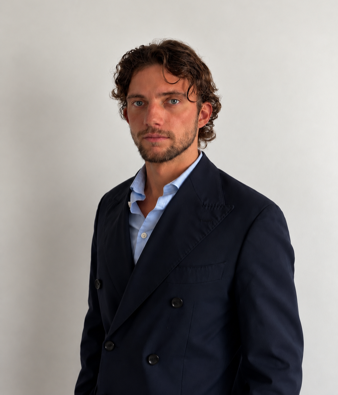 Homme avec cheveux bruns, barbe légère, portant une veste noire et une chemise bleue claire, posé contre un mur blanc.
