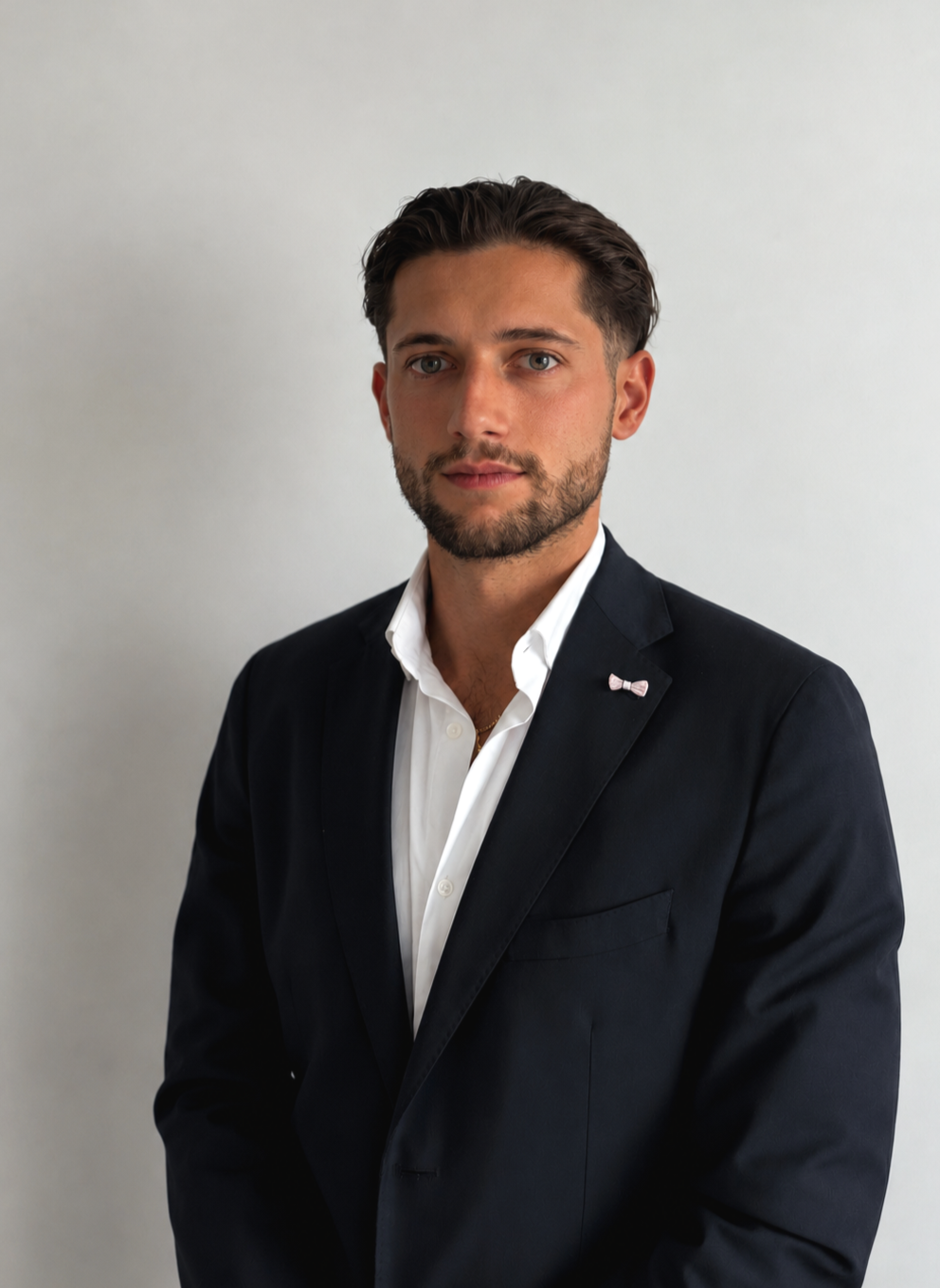 Portrait d'un homme en costume noir avec chemise blanche, debout contre un mur blanc.