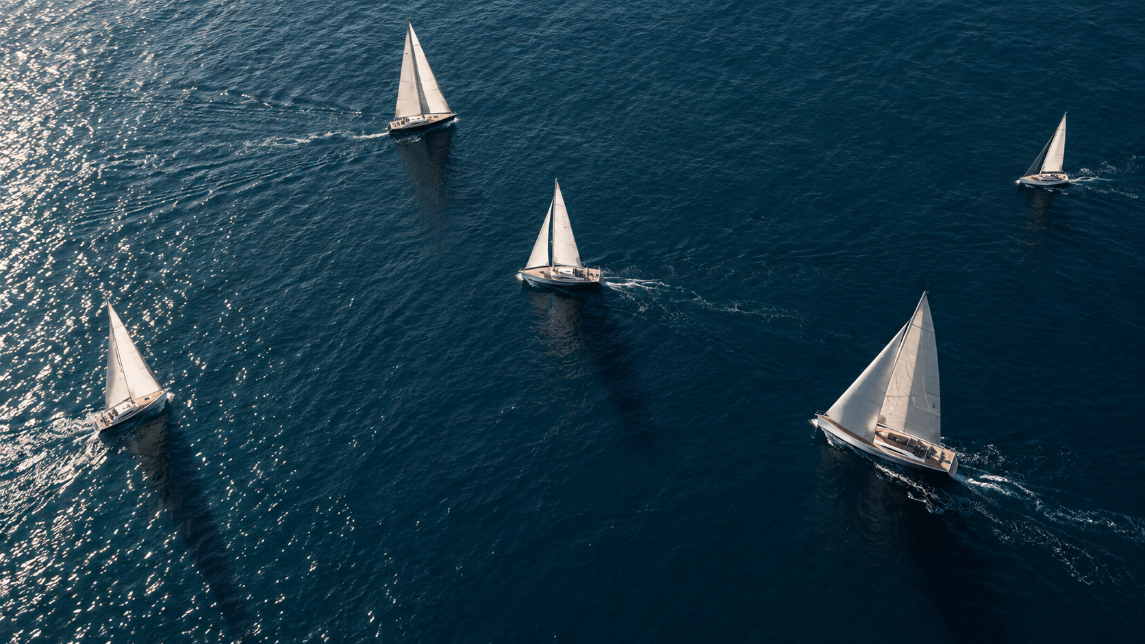 Cinq voiliers naviguant en mer, vue depuis en haut, avec l'eau bleue et le reflet du soleil.