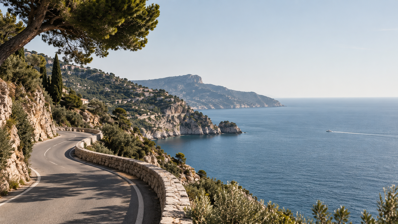 Route sinueuse longeant la mer, avec une falaise escarpée bordée de végétation et de maisons, vue sur la côte méditerranéenne sous un ciel bleu clair.