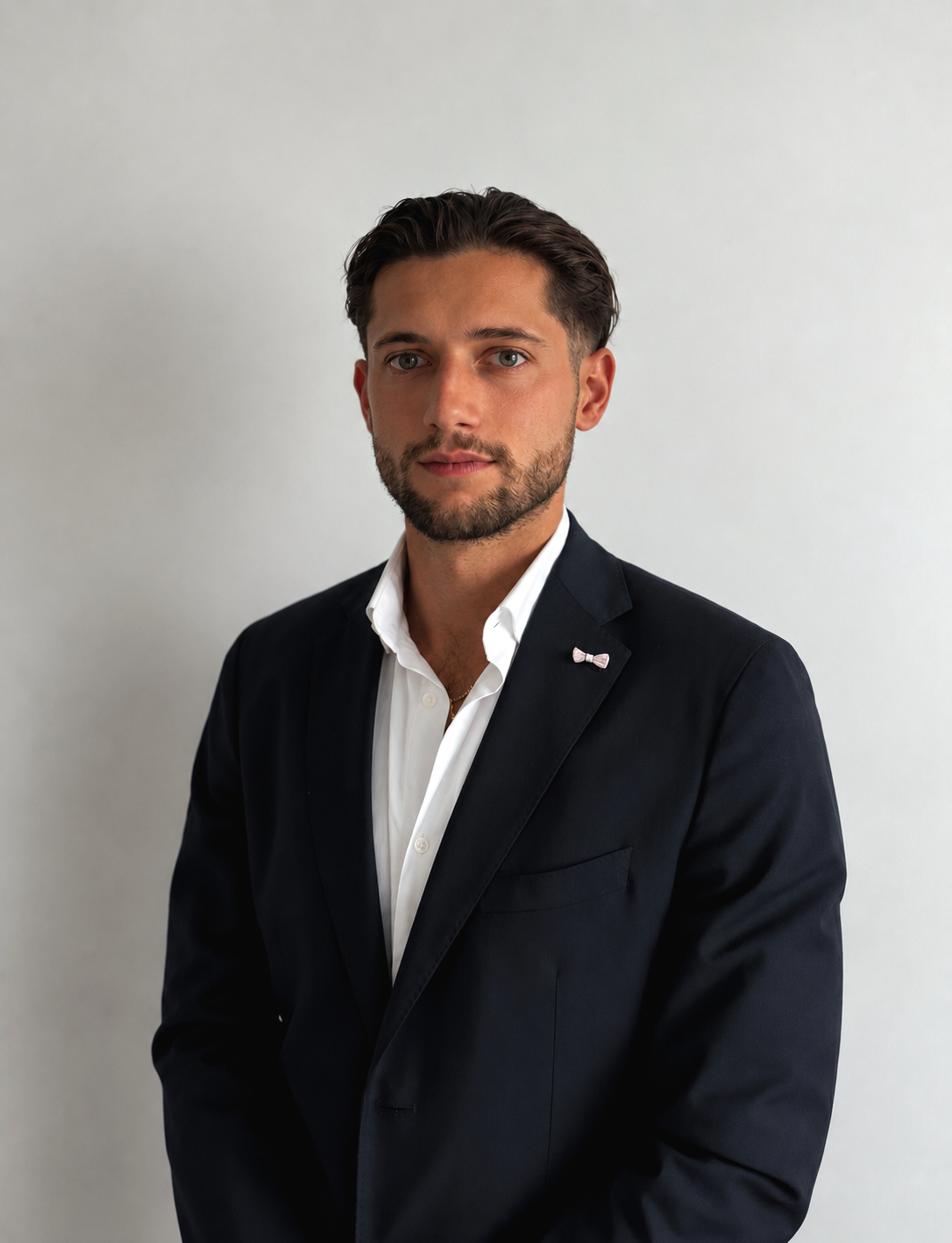 Un homme portant un blazer noir et une chemise blanche, posant devant un mur blanc.