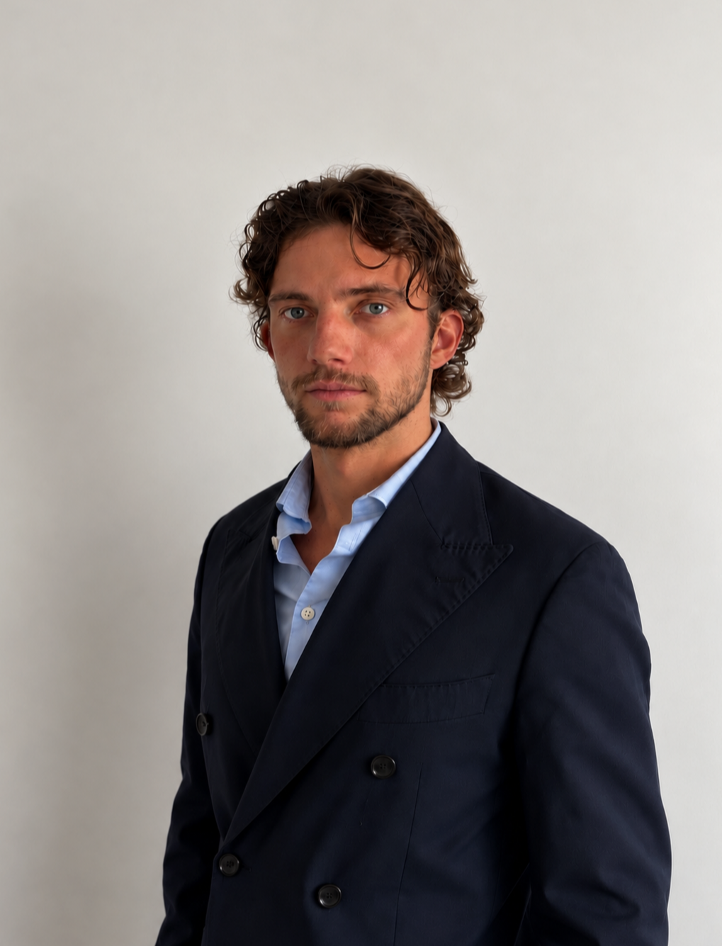 Jeune homme aux cheveux bouclés, portant un blazer noir et une chemise bleue claire, posant contre un fond blanc