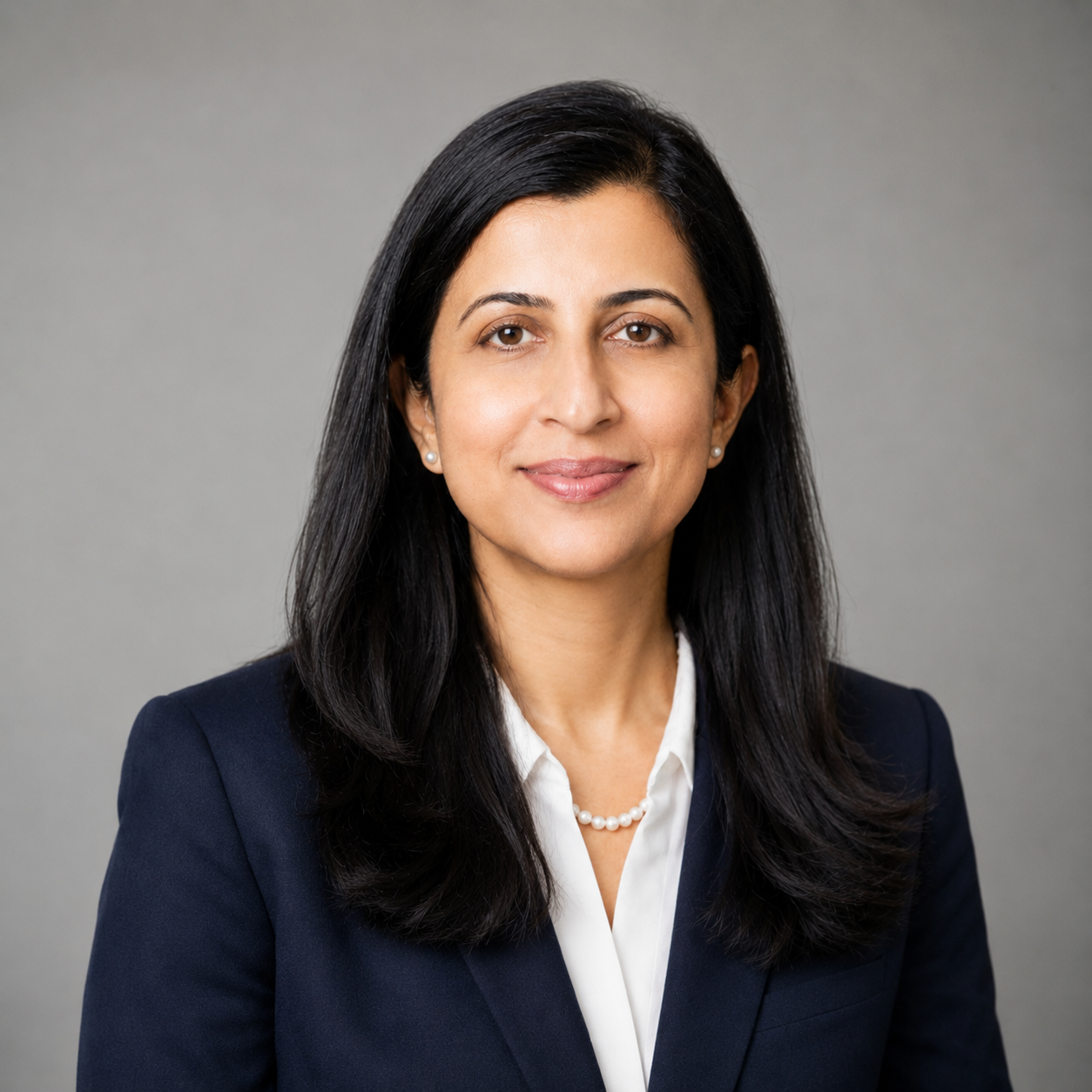 Professional headshot of a woman with long black hair, wearing a navy blazer, white blouse, and pearl earrings, smiling slightly against a gray background.