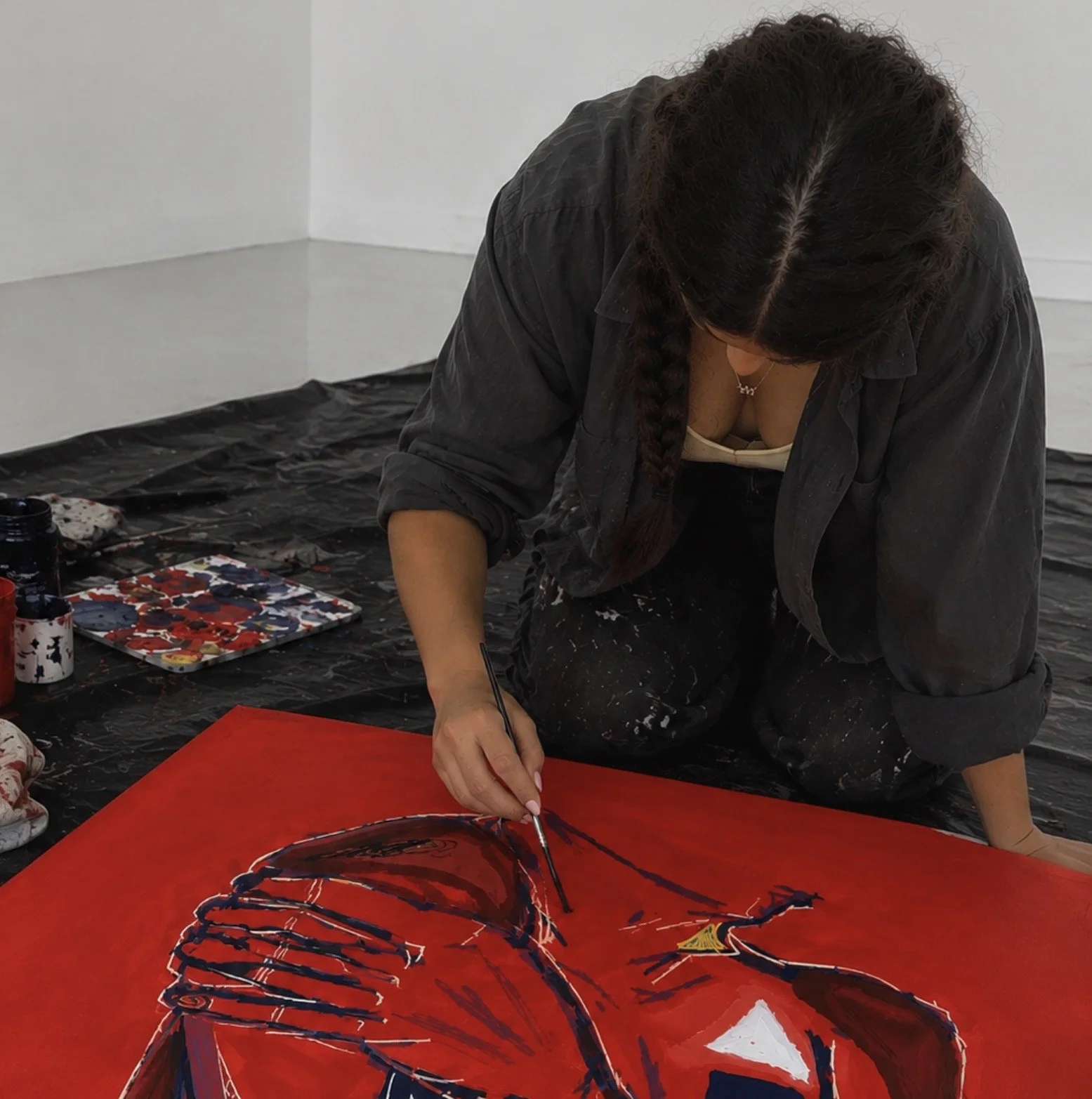 An artist creating a vibrant abstract painting on a red canvas, sitting on the floor with art supplies around, in an indoor space.
