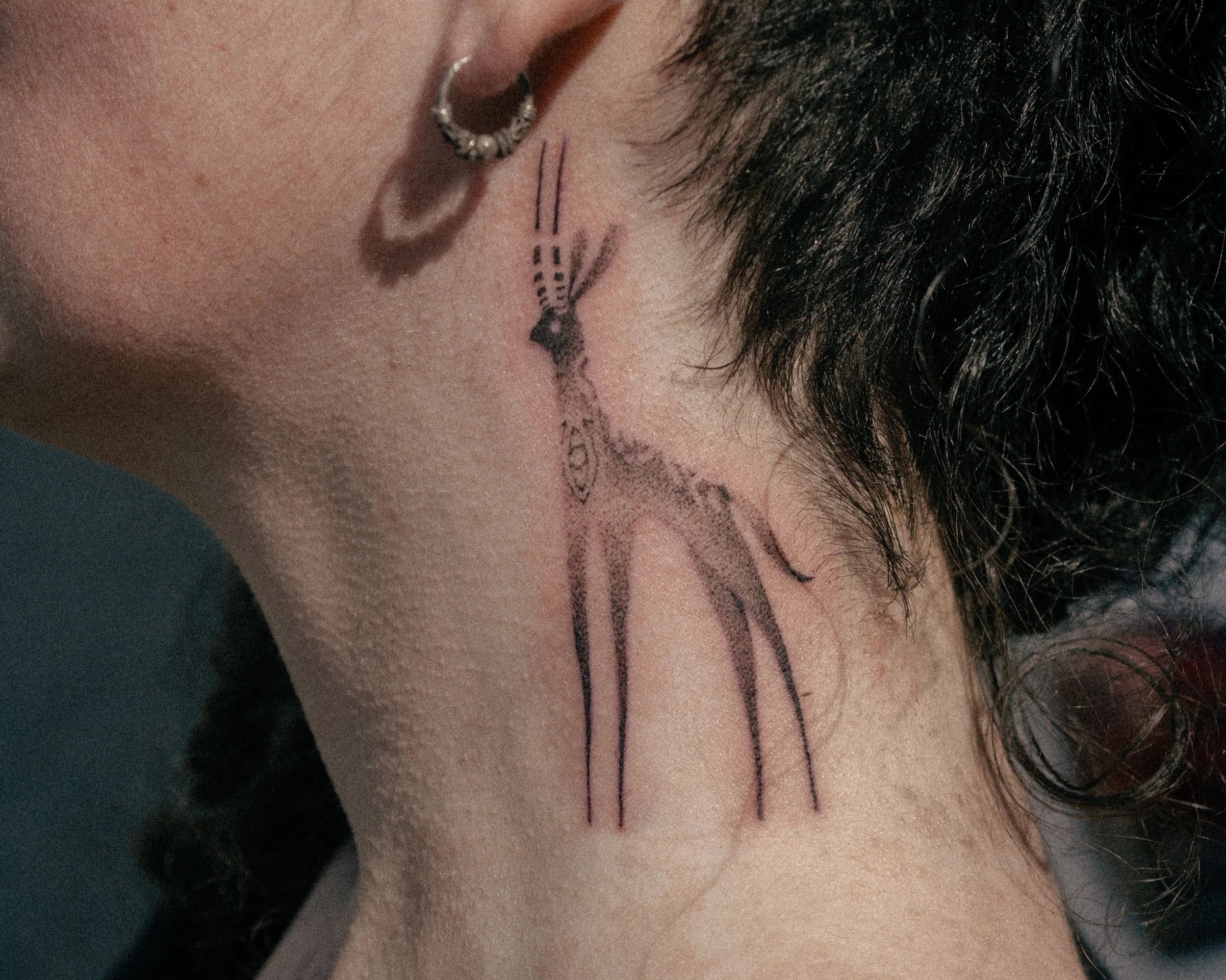 Tatouage d'une girafe sur la nuque d'une personne, avec une boucle d'oreille en perles à la narine et des cheveux noirs courts.