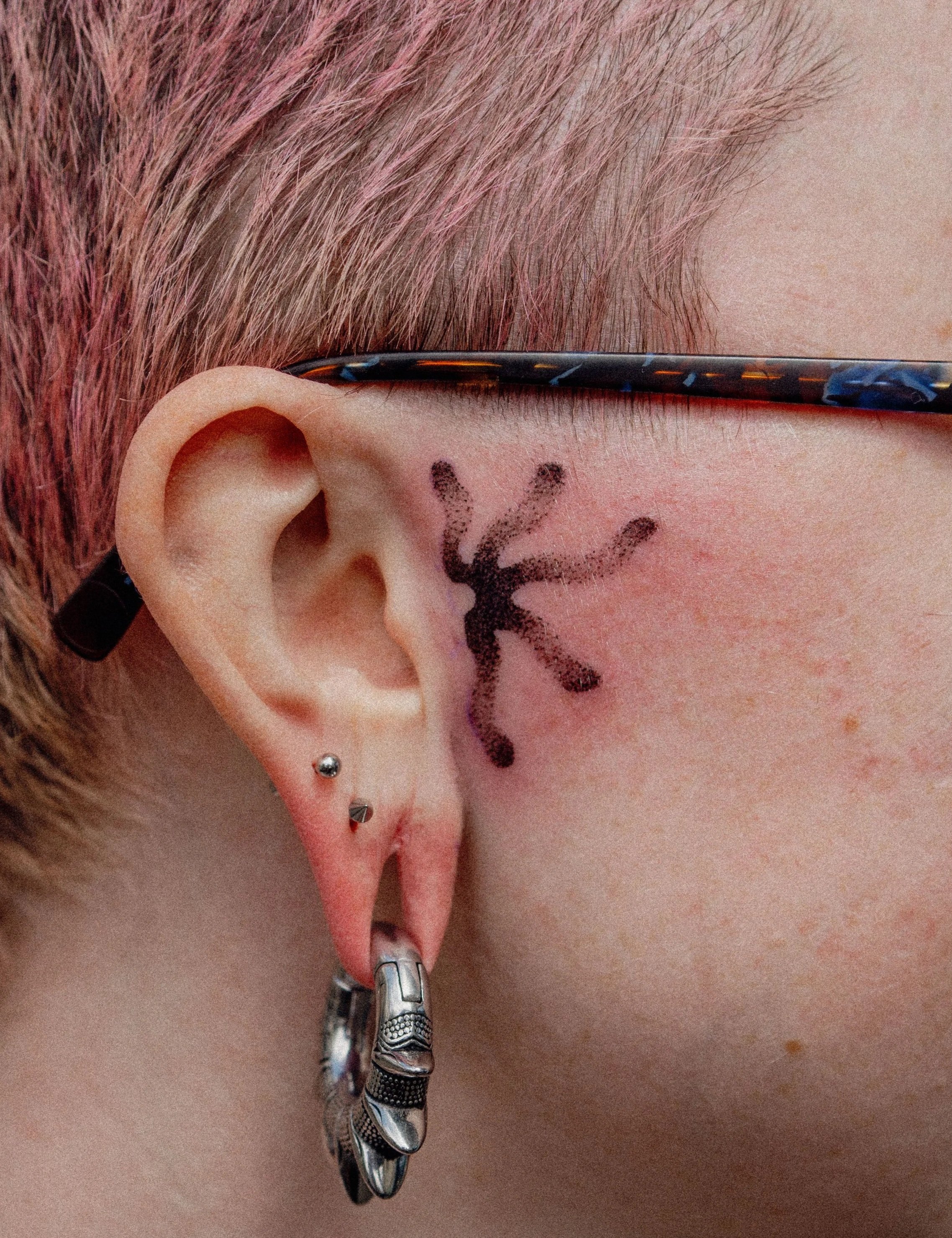 Détail d'une oreille avec plusieurs piercings, tatouage noir derrière l'oreille, lunettes avec monture colorée, et doigt avec un anneau en métal chargé d'une aiguille ou d'un outil.