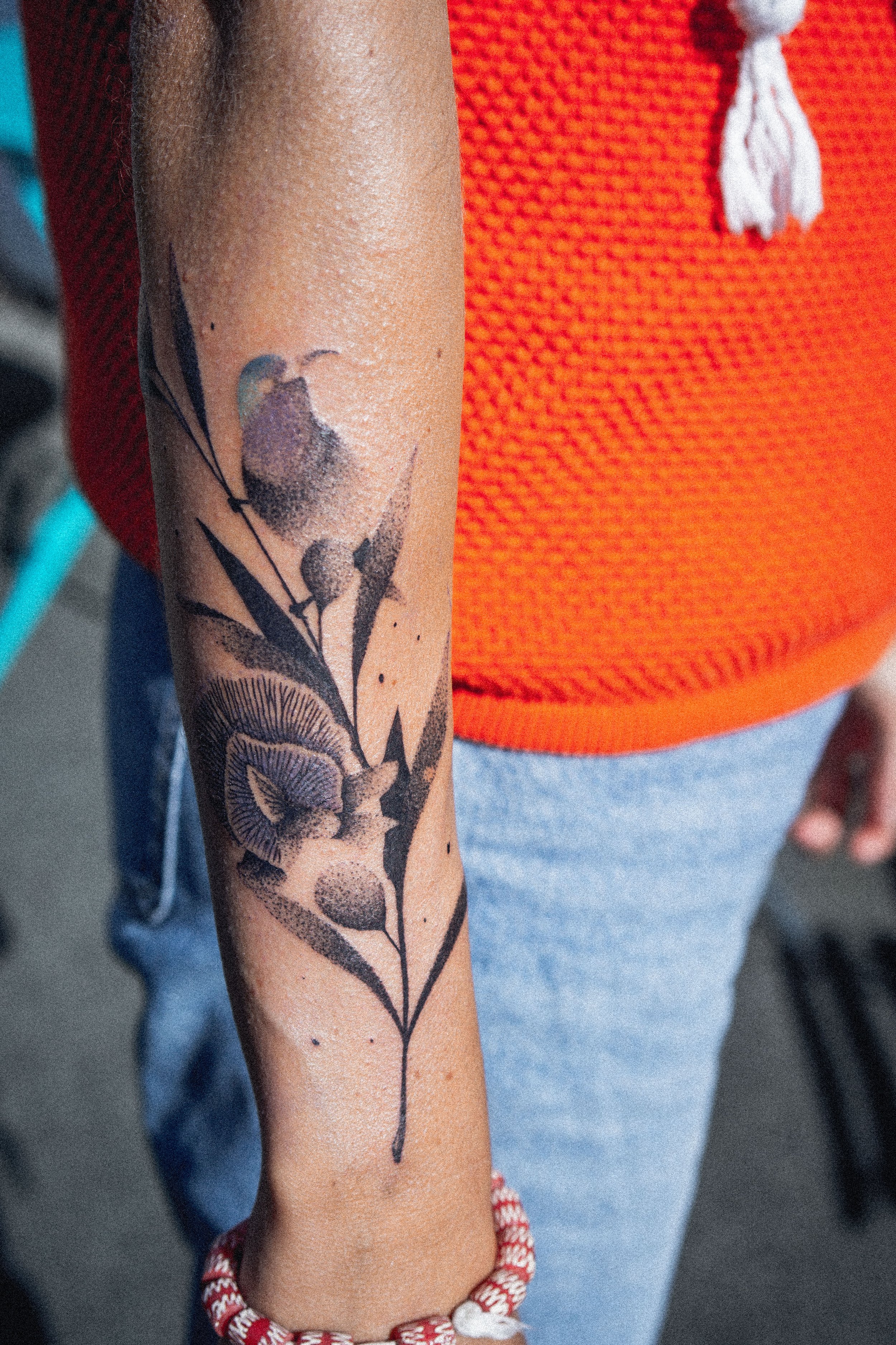 Tatouage d'une fleur sur le bras d'une personne portant un bracelet rouge et blanc, vêtue d'un pull orange et de jeans.