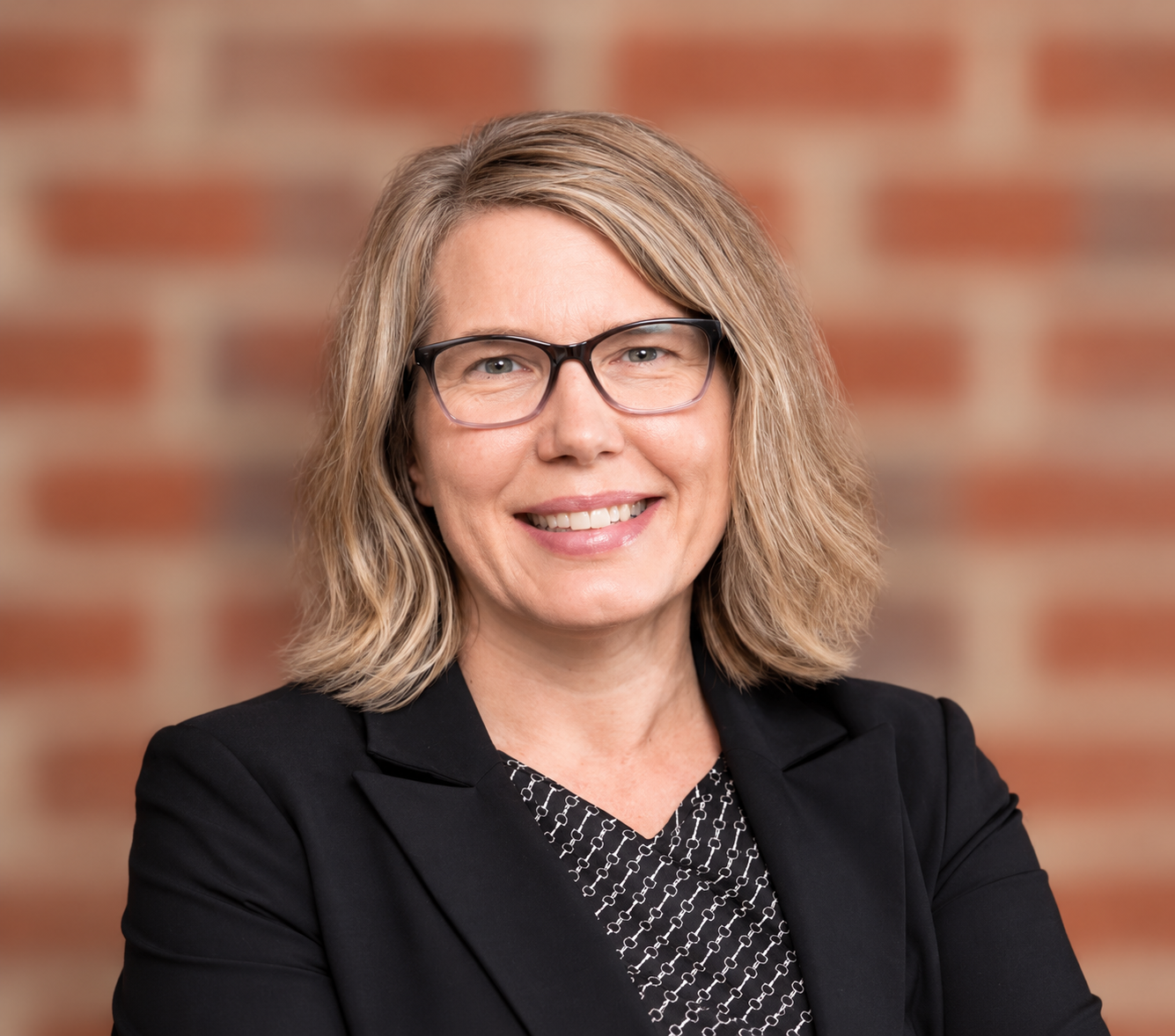 Headshot of Dr. Karie Gibson,  a smiling woman with blonde hair, wearing glasses and a black blazer, against a brick wall background.