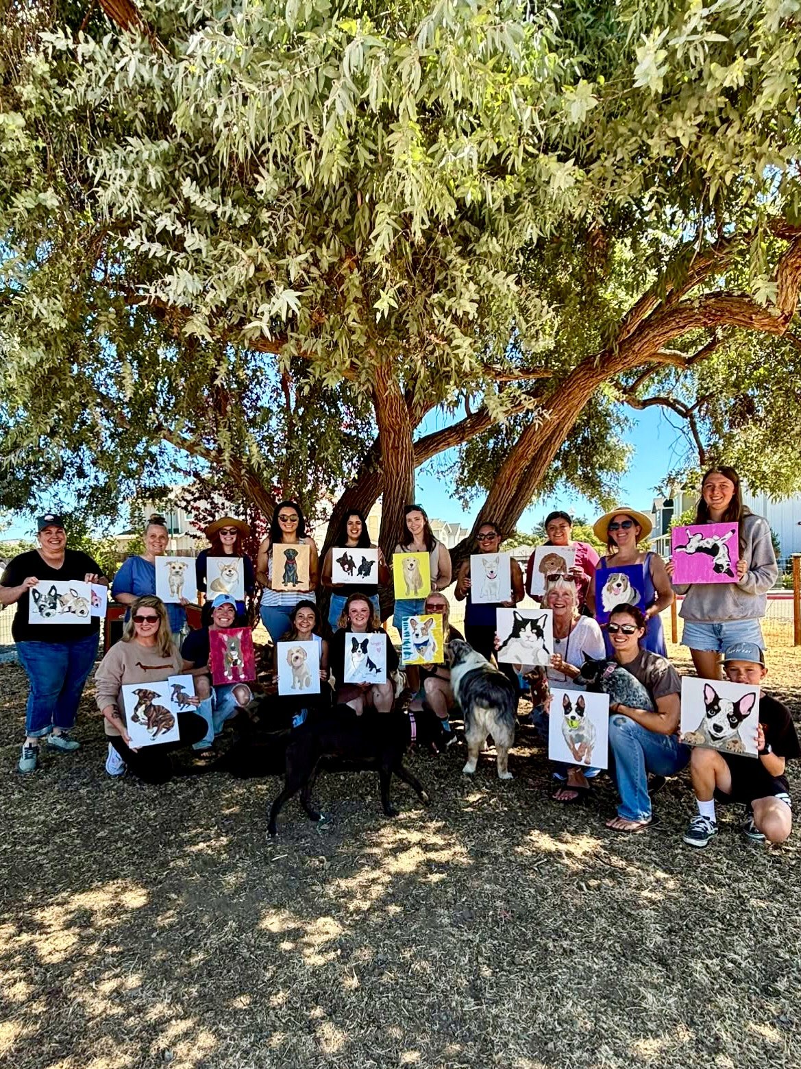 A group of women posing outdoors beneath a large leafy tree, smiling & holding colorful paintings of dogs at a dog-themed painting event with several dogs gathered around them