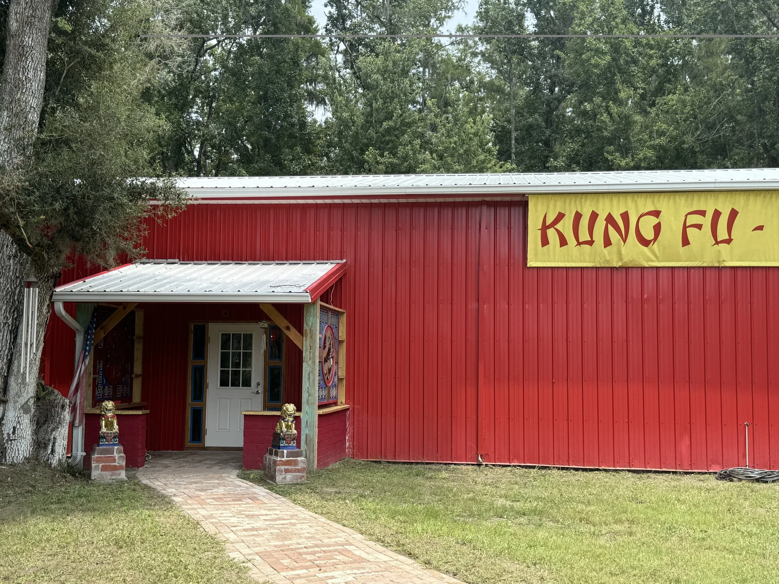 Red building with a yellow martial arts banner that has red characters, surrounded by trees and a brick pathway leading to the entrance, decorated with two Chinese lion statues.