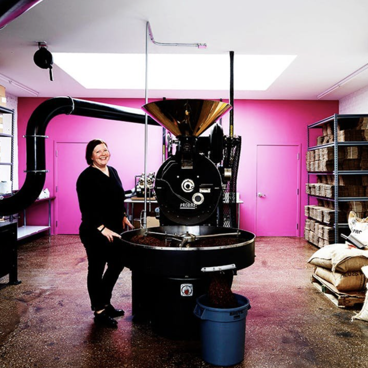 Person in a coffee roasting facility with pink walls, standing beside a large coffee roaster, surrounded by bags and shelves of coffee beans.