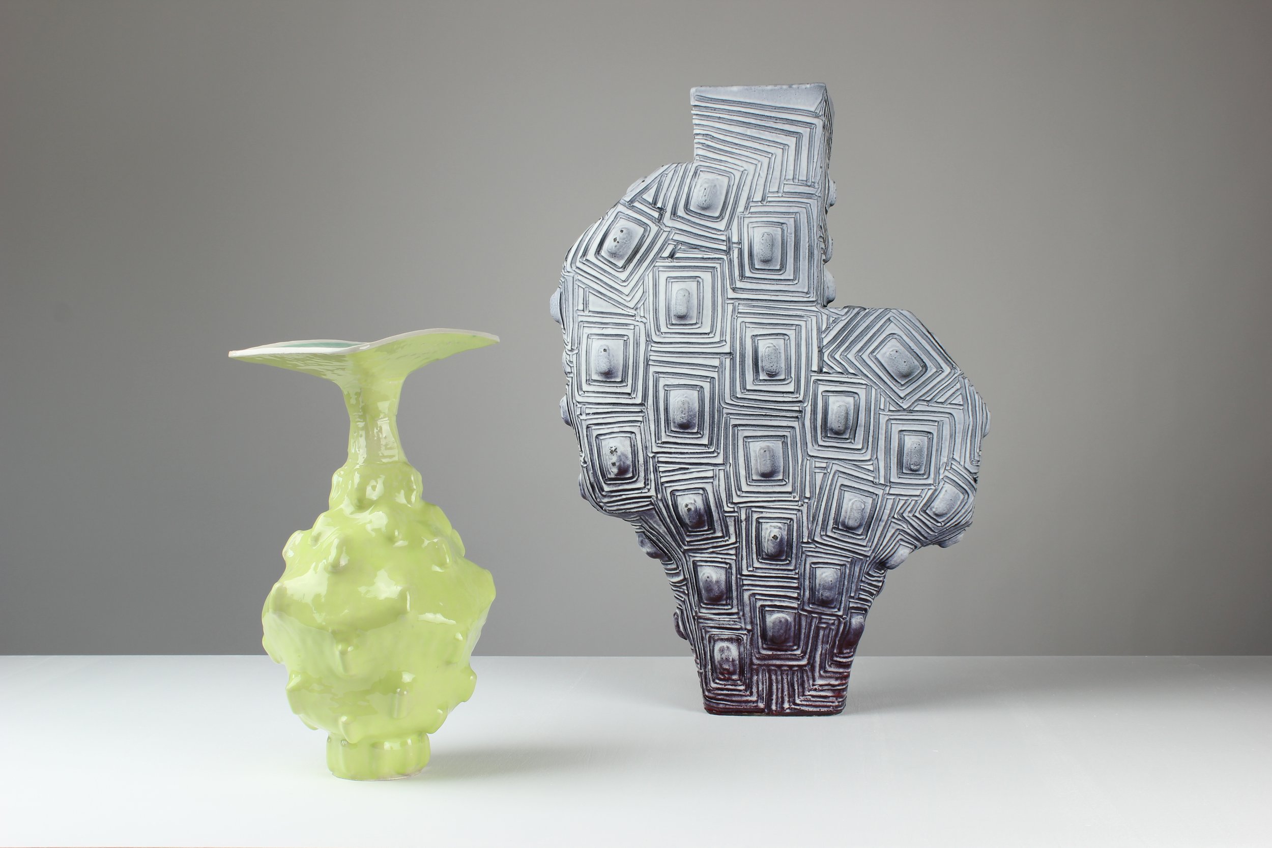 Drei skulpturale Keramikvasen, eine in leuchtendem Gelb mit unregelmäßiger Oberfläche, eine in Grau und Schwarz mit geometrischem Muster, und eine kleine grüne Vase, alle auf einer weißen Oberfläche gegen einen grauen Hintergrund.