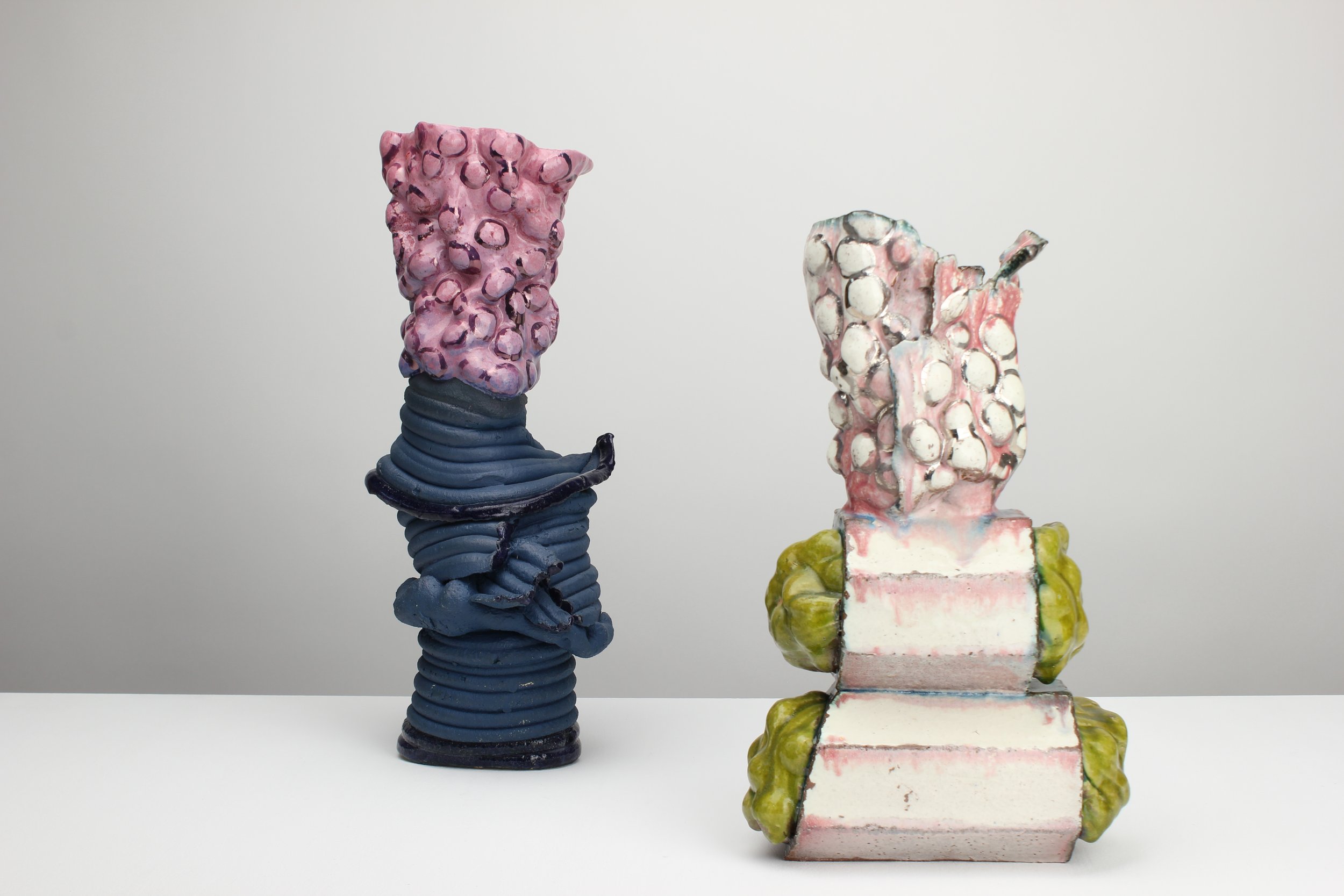 Kunstvolle Skulpturen aus buntem Keramikmaterial, zwei Figuren mit welliger Textur und vielfältigen Farben, auf einer weißen Oberfläche vor einem grauen Hintergrund.