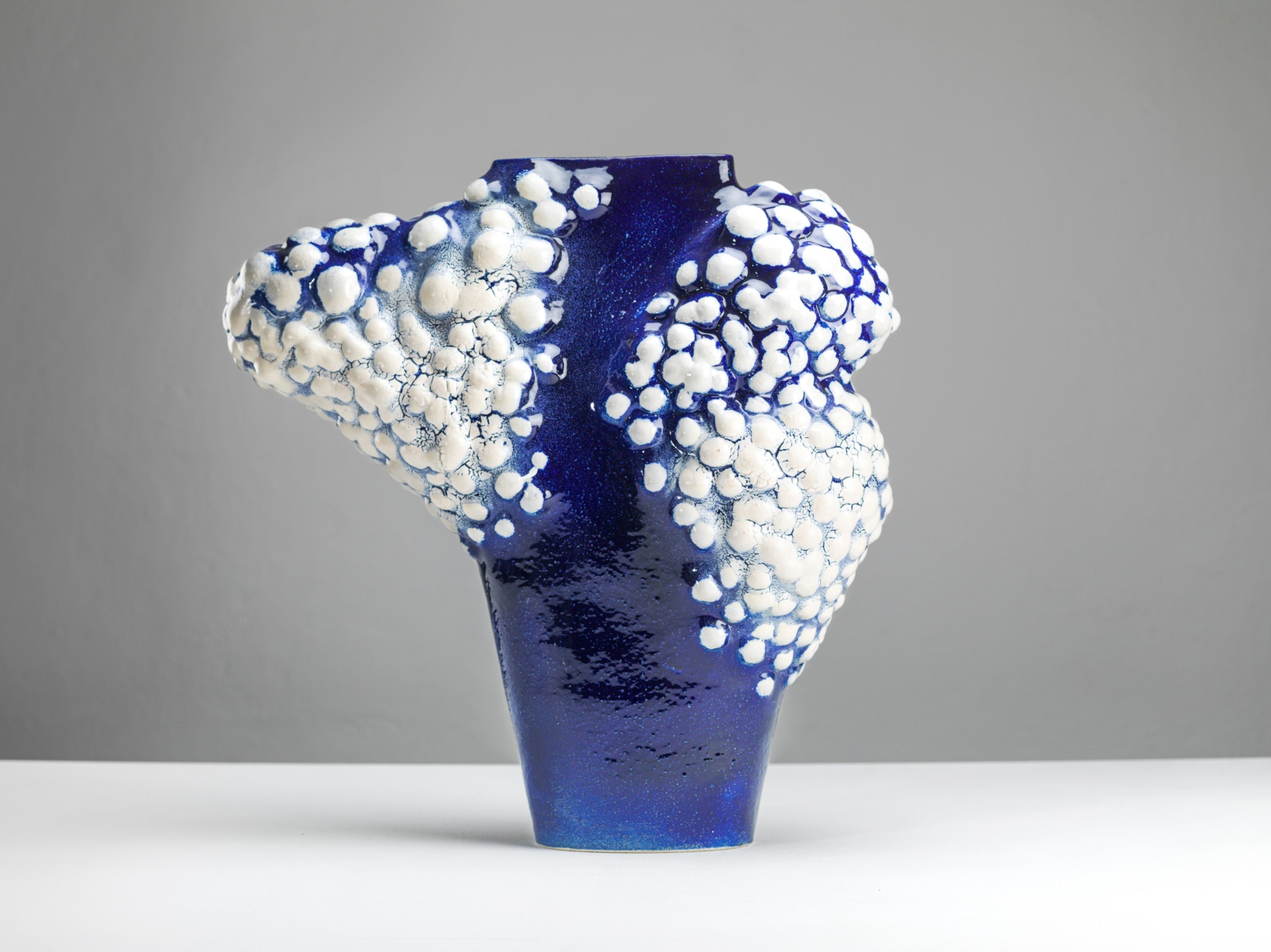 Eine blaue Glaskunst-Vase mit weißen, kugelartigen Mustern und bläulichen, netzartiger Strukturen auf der Oberfläche, vor einem grauen Hintergrund.