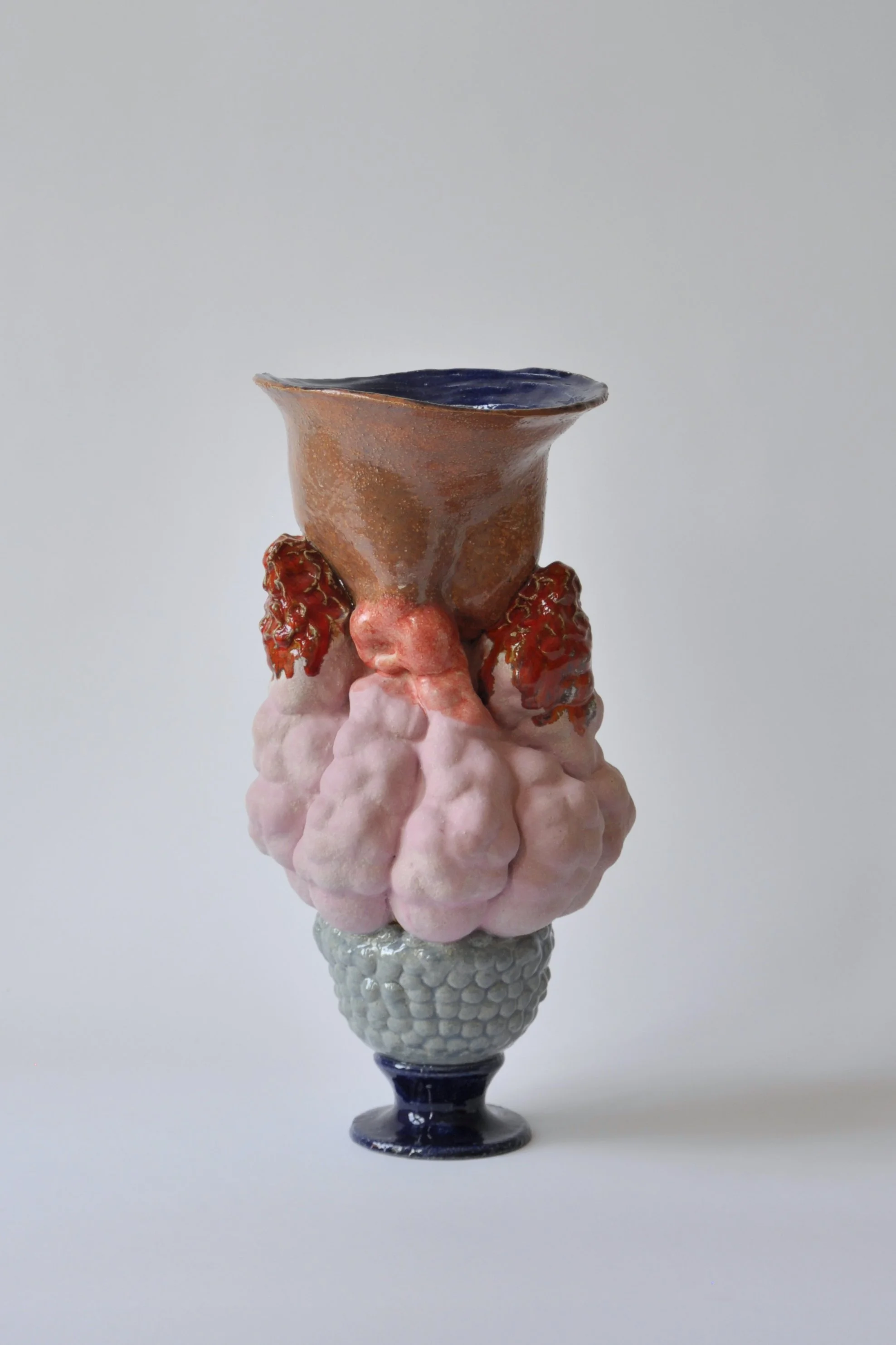 Ein kunstvoller Keramikbecher mit rosa Wolken und roten Felsen, auf einem blauen Sockel, vor einem weißen Hintergrund.