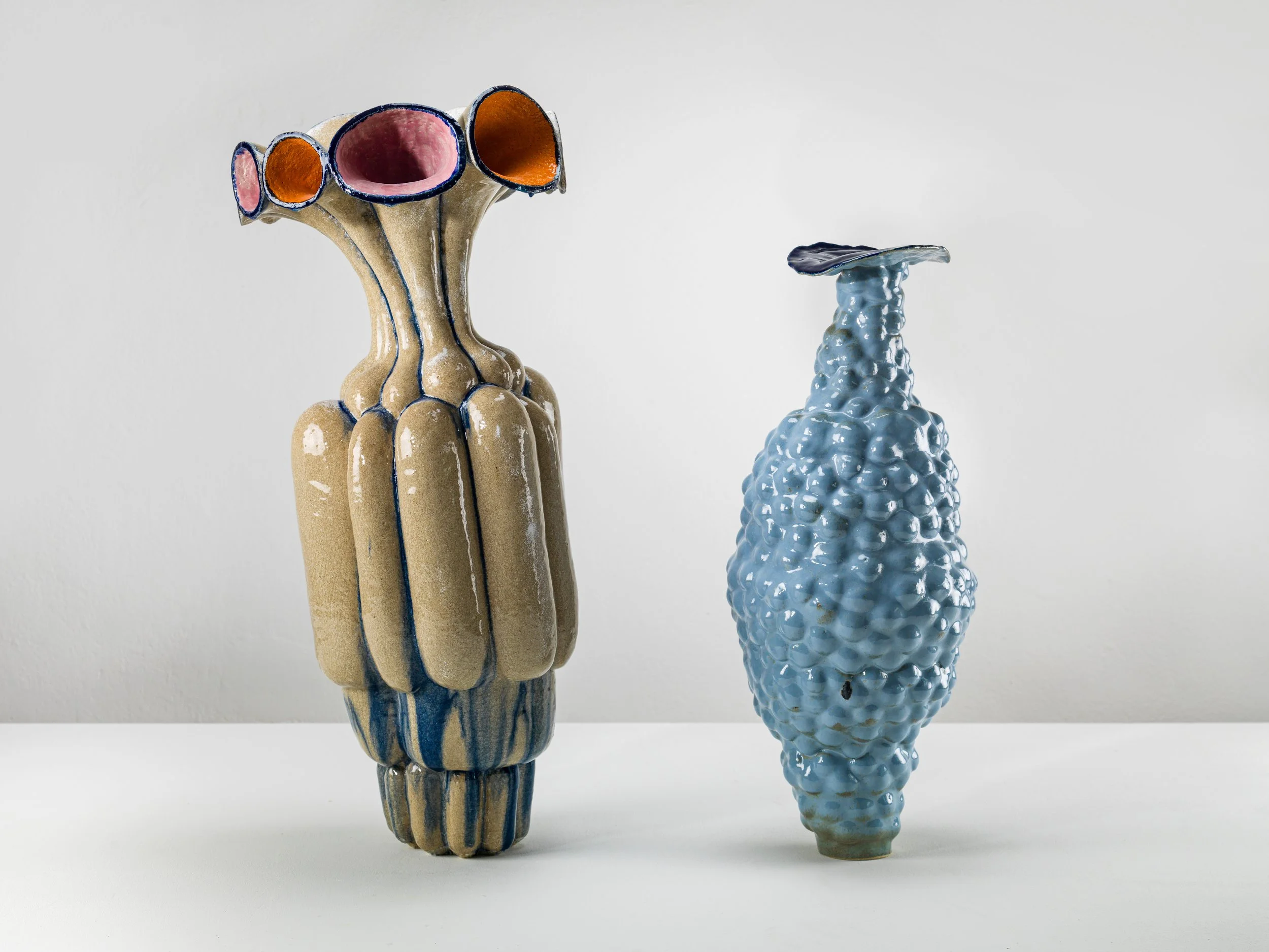 Zwei skulpturale Keramikvasen, eine bauchig und mit Farbglasur in beige, rosa und orange, die andere schlanker und bläulich mit bügelartiger Oberseite, vor weißem Hintergrund.