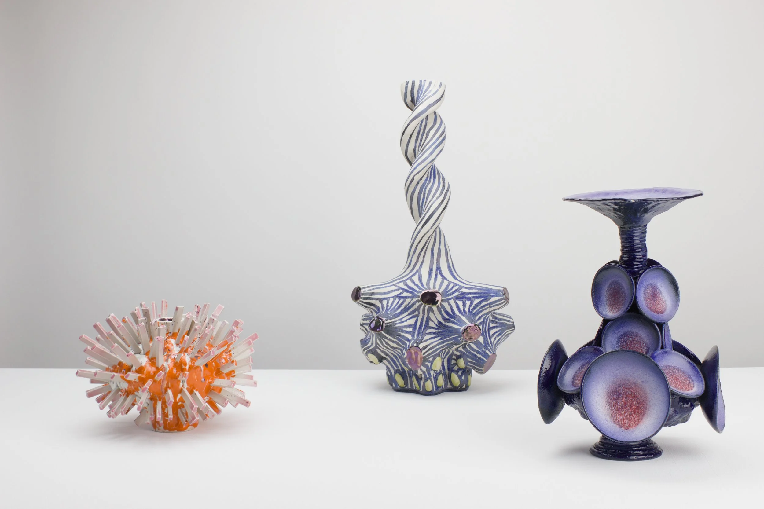Kunstvolle Keramik- und Kunstobjekte auf weißem Hintergrund, darunter eine orangefarbene Objekt mit rohrähnlichen Strukturen, eine blau-weiße gedrehte Skulptur, eine blaue Keramik mit blumenähnlichem Design und eine violette Skulptur.