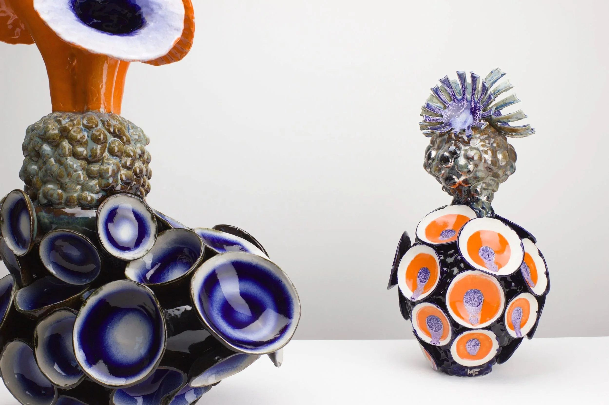 Bunte Glas-Objekte, die wie Blumen oder Pilze geformt sind, mit bunten Mustern in Blau, Weiß, Orange und Lila auf weißem Hintergrund.