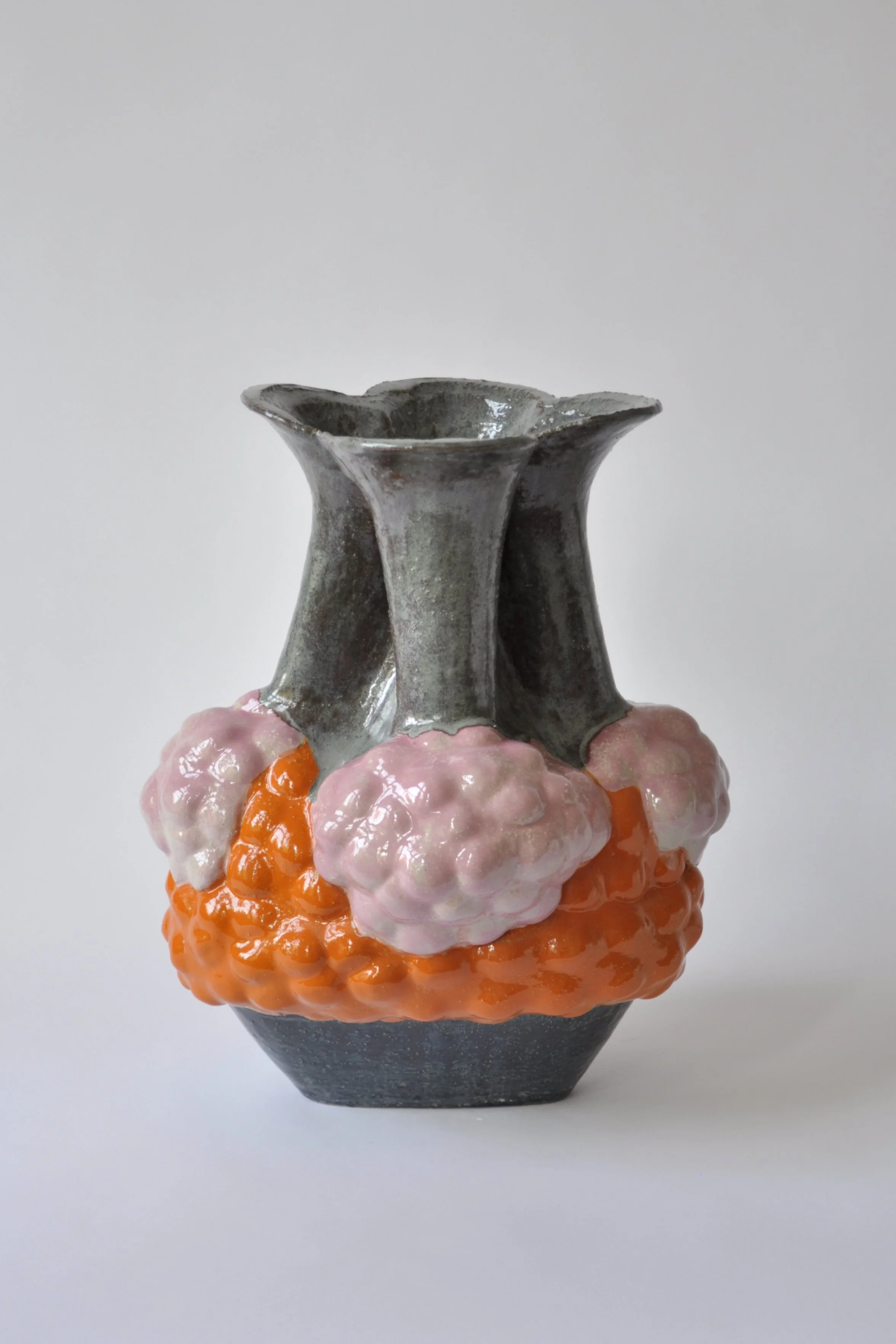 Eine keramikartige Vase in grau mit einer offenen, unregelmäßig geformten Oberseite, die auf einem Sockel mit einer Schicht aus pinkfarbenem und orangefarbenem Gelee oder Modellage steht, vor einem weißen Hintergrund.