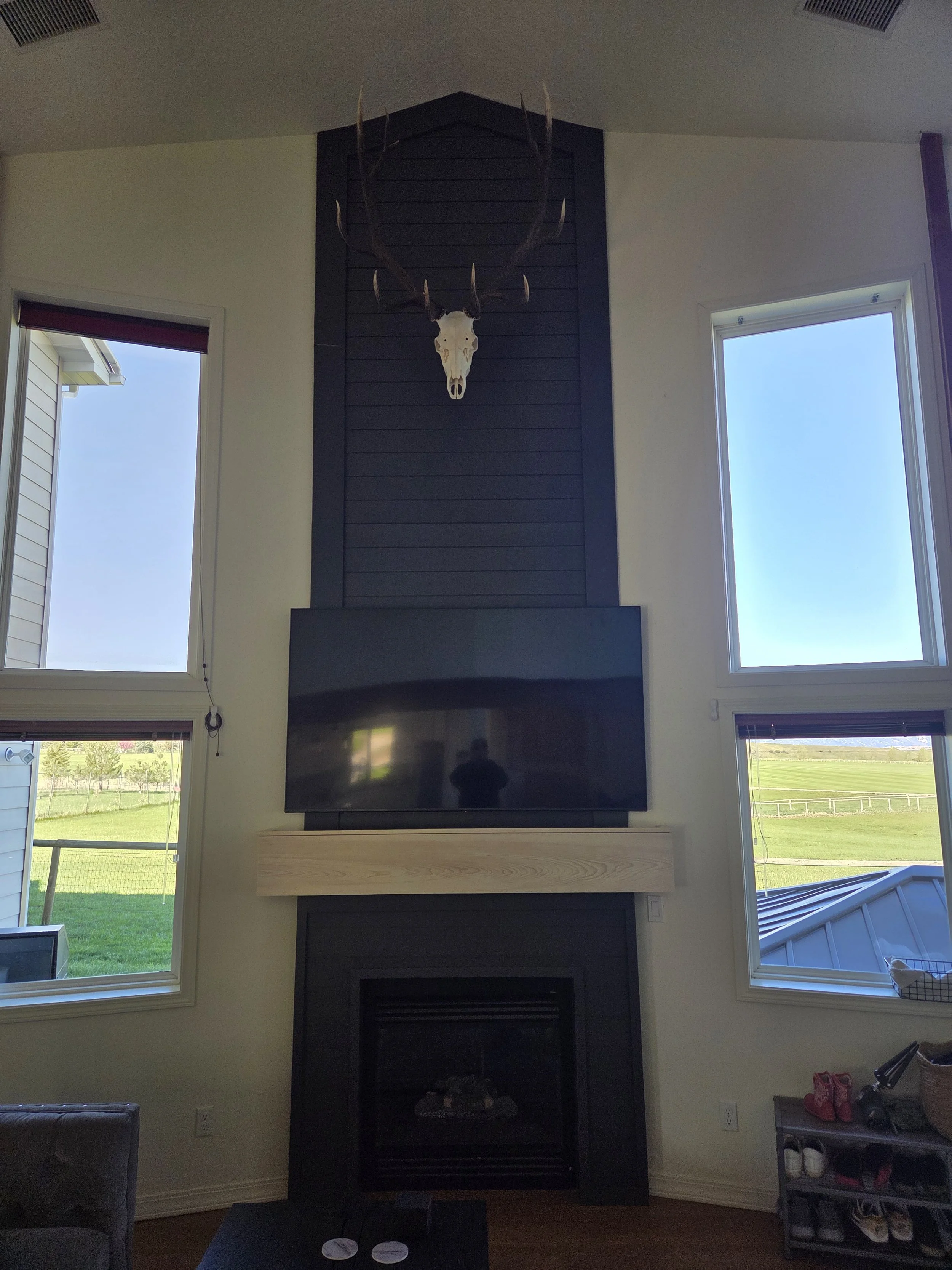 Living room interior with three large windows showing a green field and blue sky outside, a black fireplace with a mounted animal skull with antlers, a wooden shelf below the fireplace, and a small shelving unit with shoes and red and black items on the right side.