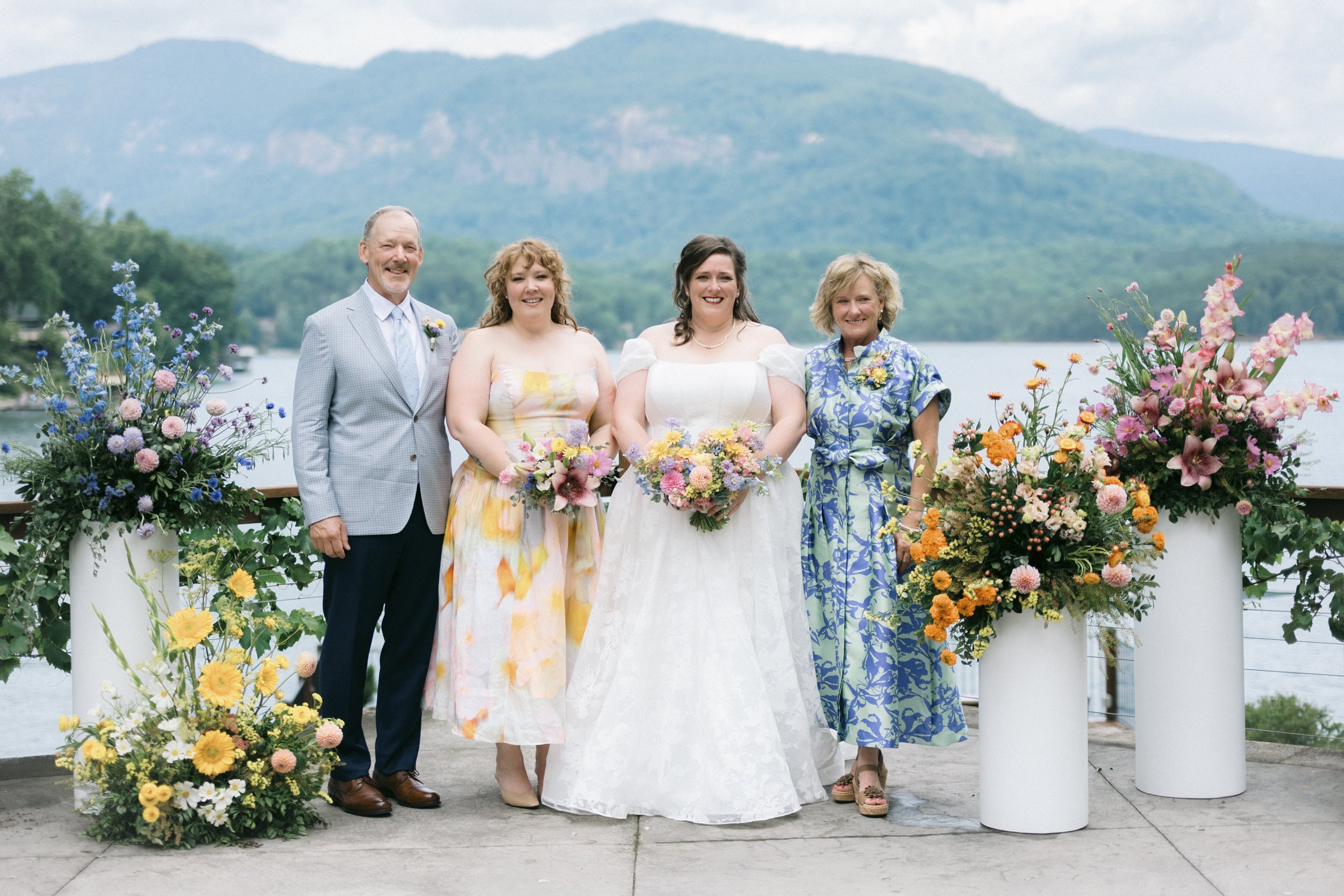 A bride and her three wedding guests stand on an outdoor dock near a lake, with mountains in the background, all smiling and holding bouquets, surrounded by colorful flowers.