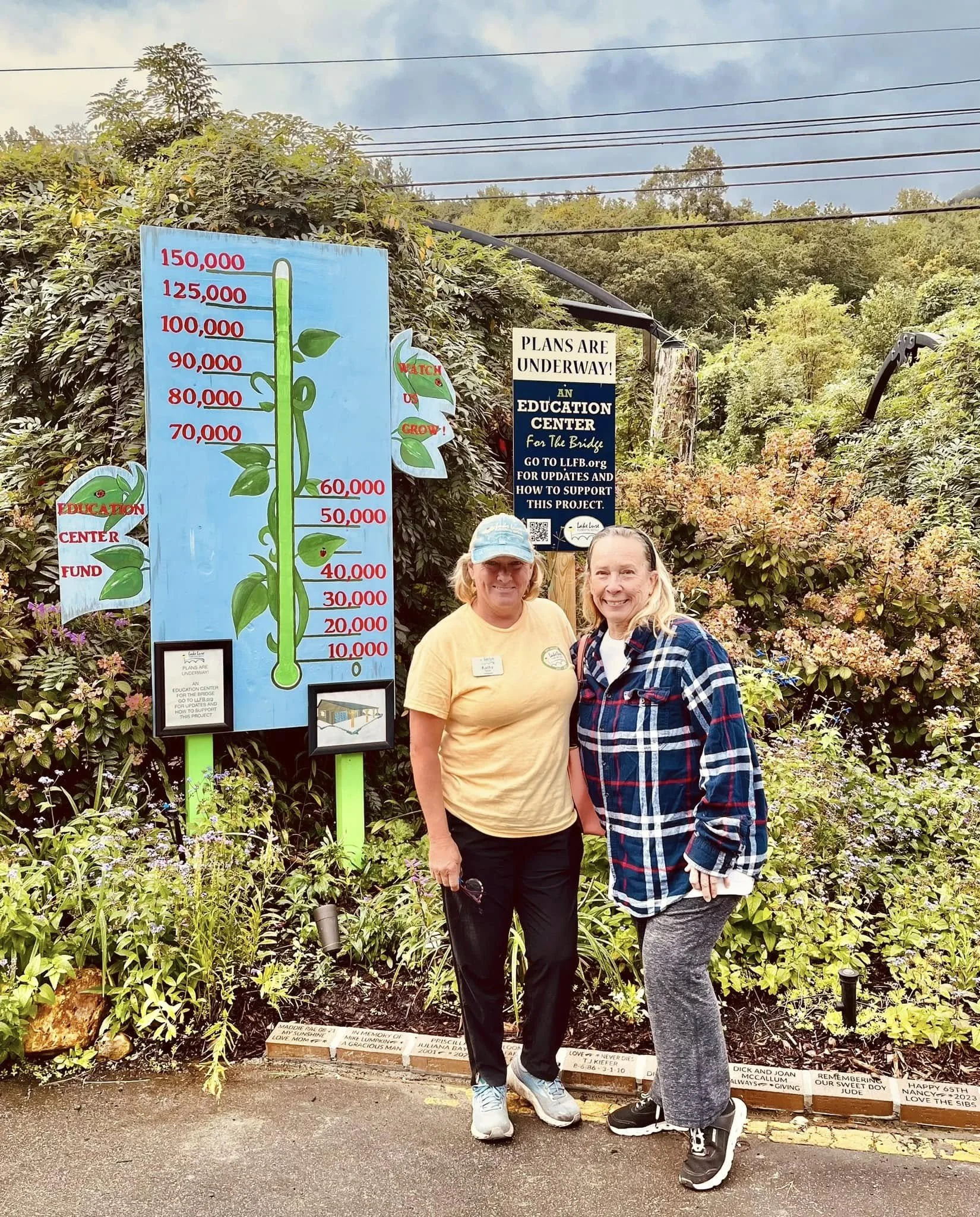 Two women standing in front of a memorial wall with flowers and inscriptions. One woman is wearing a yellow shirt and baseball cap, and the other is wearing a plaid shirt and gray leggings. Behind them, there are signs including a tall plant-themed fundraiser thermometer and a sign that reads, 'Plans are underway! An Education Center for the Bridge, go to llfb.org for updates and how to support this project.'
