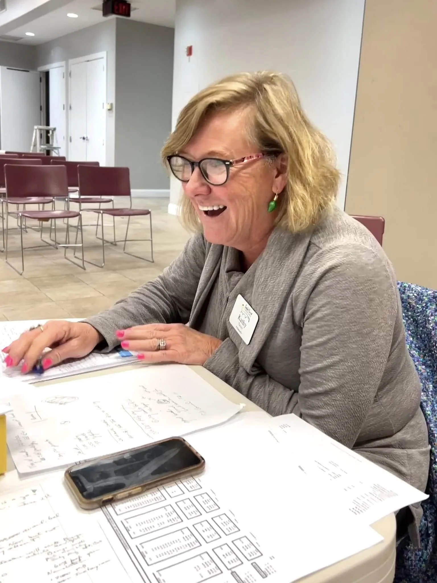 A woman with blonde hair, glasses, and green earrings is smiling and laughing while sitting at a table covered with papers, notebooks, and a smartphone. She appears to be in a meeting or workshop setting.