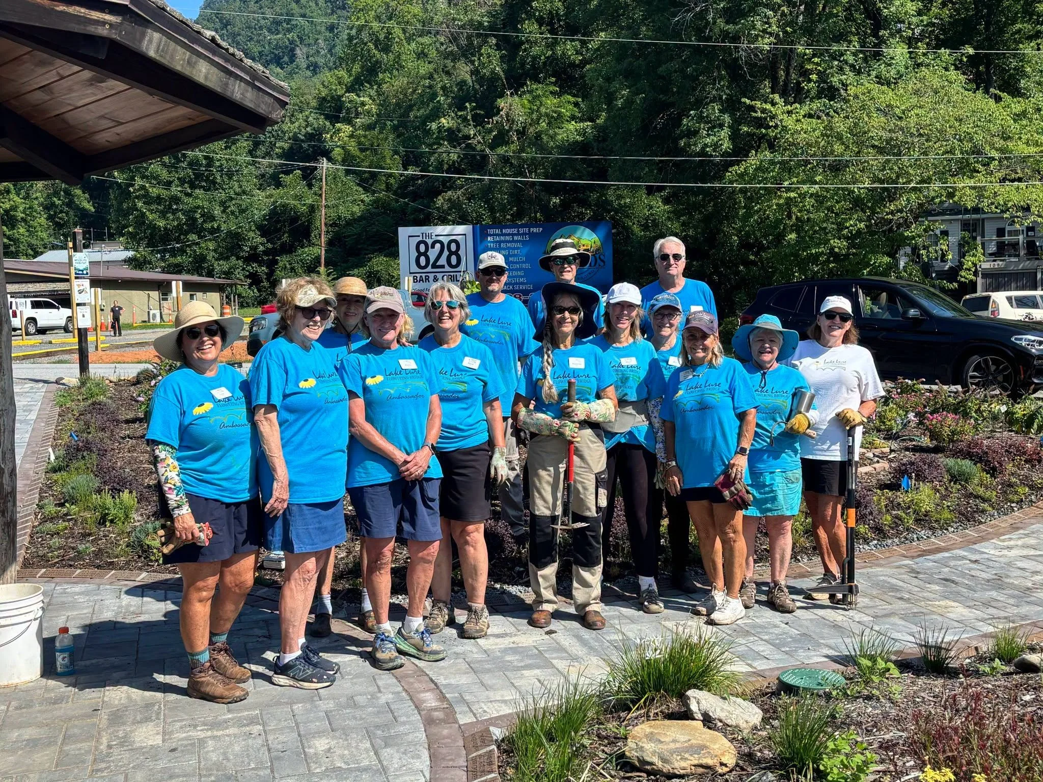 A group of approximately 15 volunteers in blue shirts standing outside in front of trees and parked cars during daytime, posing for a photo at a community planting event.