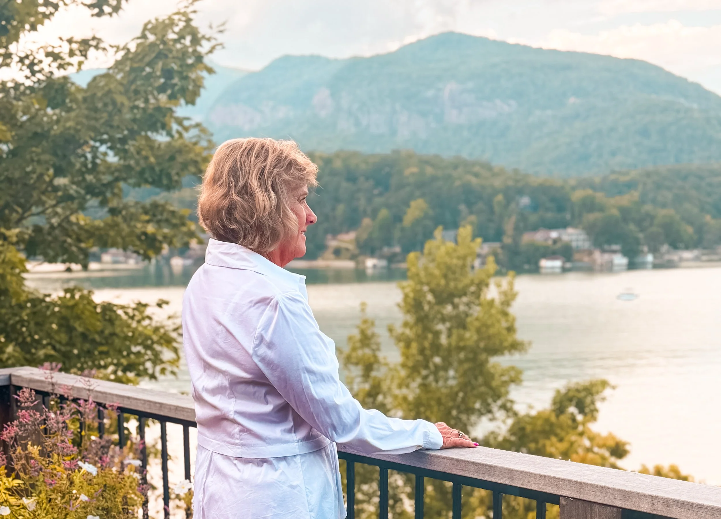 Older woman with short gray hair gazing at a scenic landscape of a lake and mountains, wearing a white shirt and standing on a balcony with flowers and a railing.
