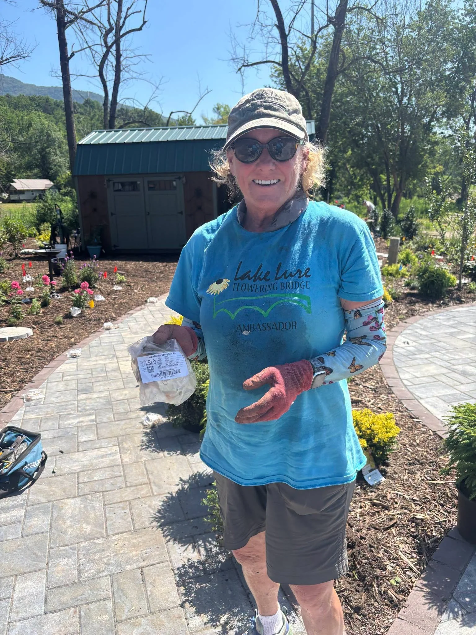 A woman wearing sunglasses, a beige cap, and a blue T-shirt smiling as she holds a bag of plant bulbs outdoors in a garden with blooming flowers and trees.