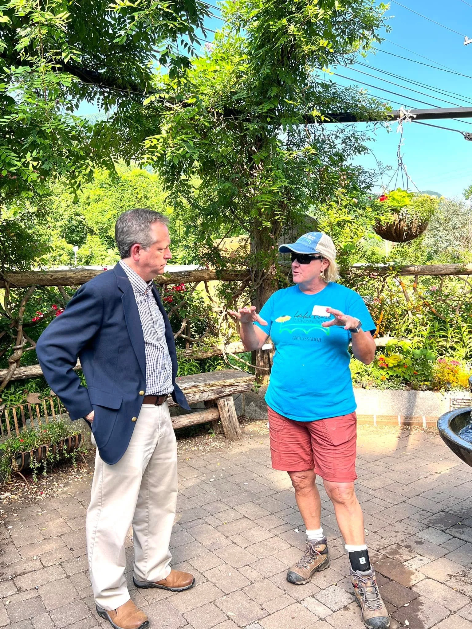 Two people standing outdoors in conversation. One is a man in a navy blazer, dress shirt, light-colored trousers, and brown shoes. The other is a woman wearing a blue T-shirt with a logo, red shorts, hiking boots, a baseball cap, and sunglasses. They are under a large tree with greenery and a hanging flower basket, with a wooden fence in the background and a bright, clear sky.