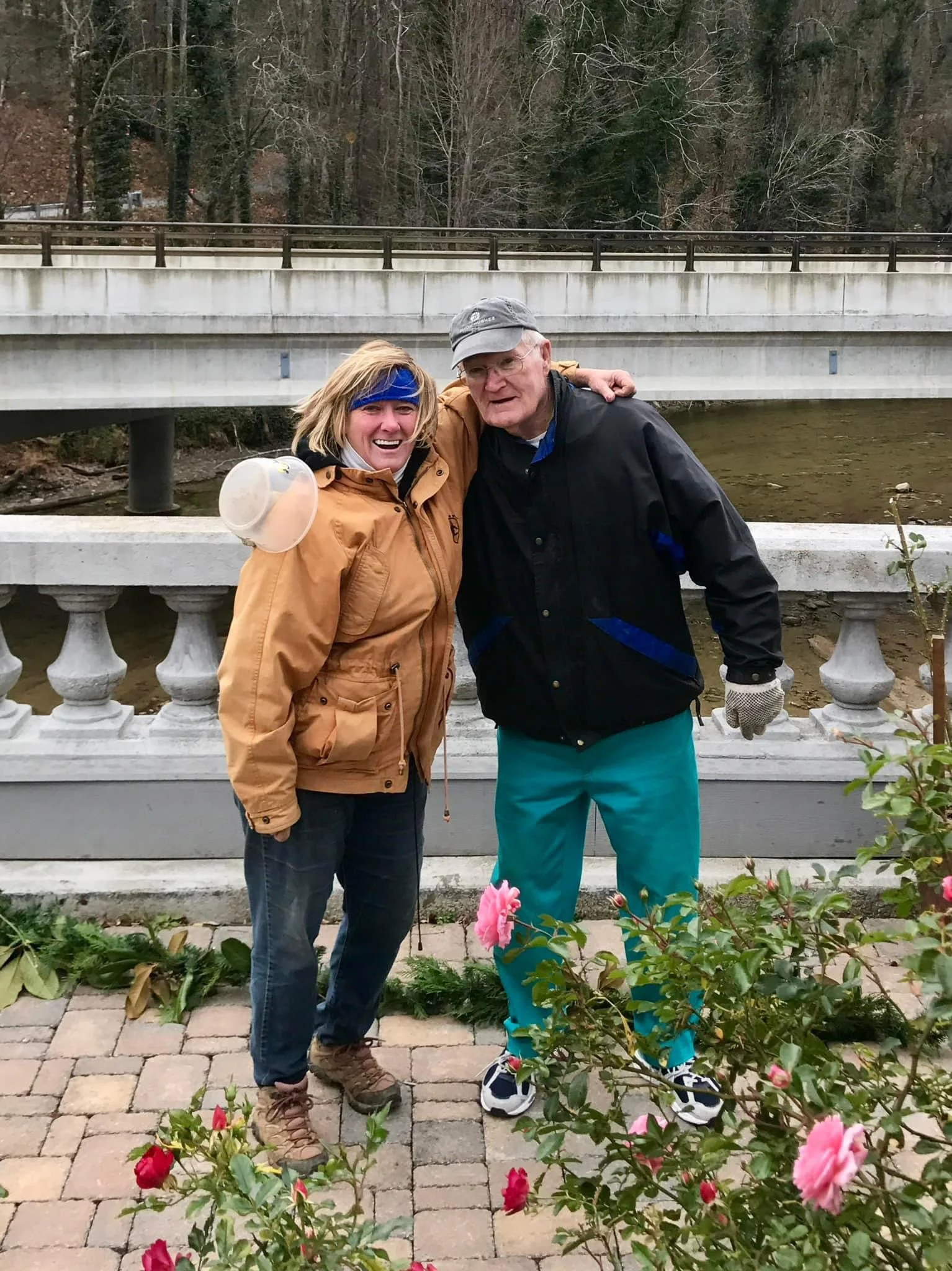 Two people smiling and standing close together outdoors on a stone-paved walkway with a bridge and trees in the background. Both are dressed warmly, and there are pink roses in the foreground.