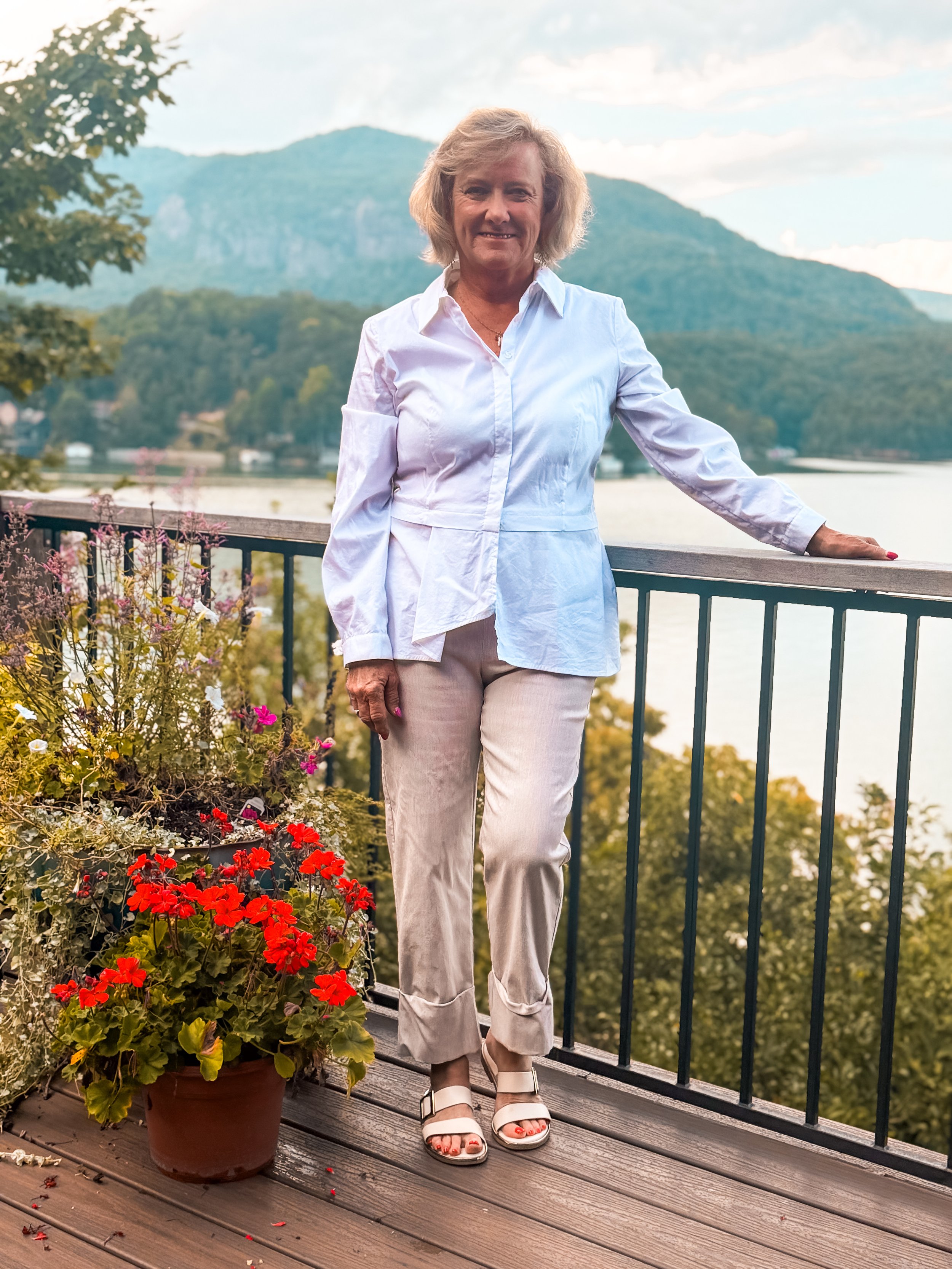 A woman standing on a wooden deck near a railing, with colorful flowers in a pot beside her, overlooking a body of water and mountains in the background, during daytime.