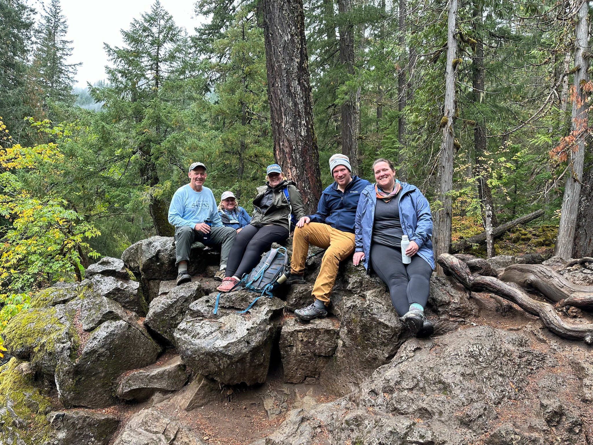 Five people sitting on rocks in a dense forest, wearing outdoor clothing and smiling.